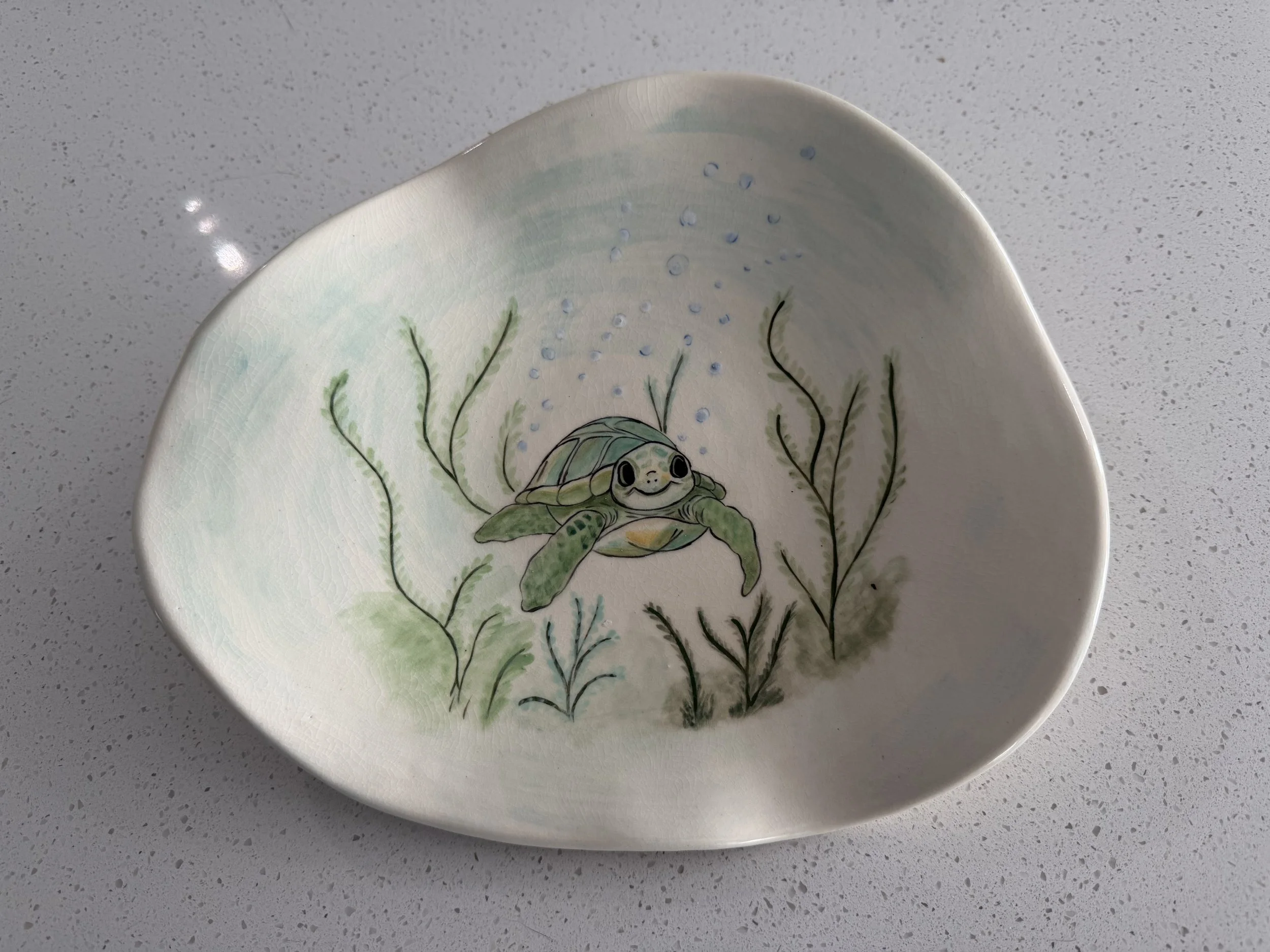 Decorative ceramic bowl with a painting of a happy turtle swimming underwater among seaweed and bubbles, on a white speckled surface.