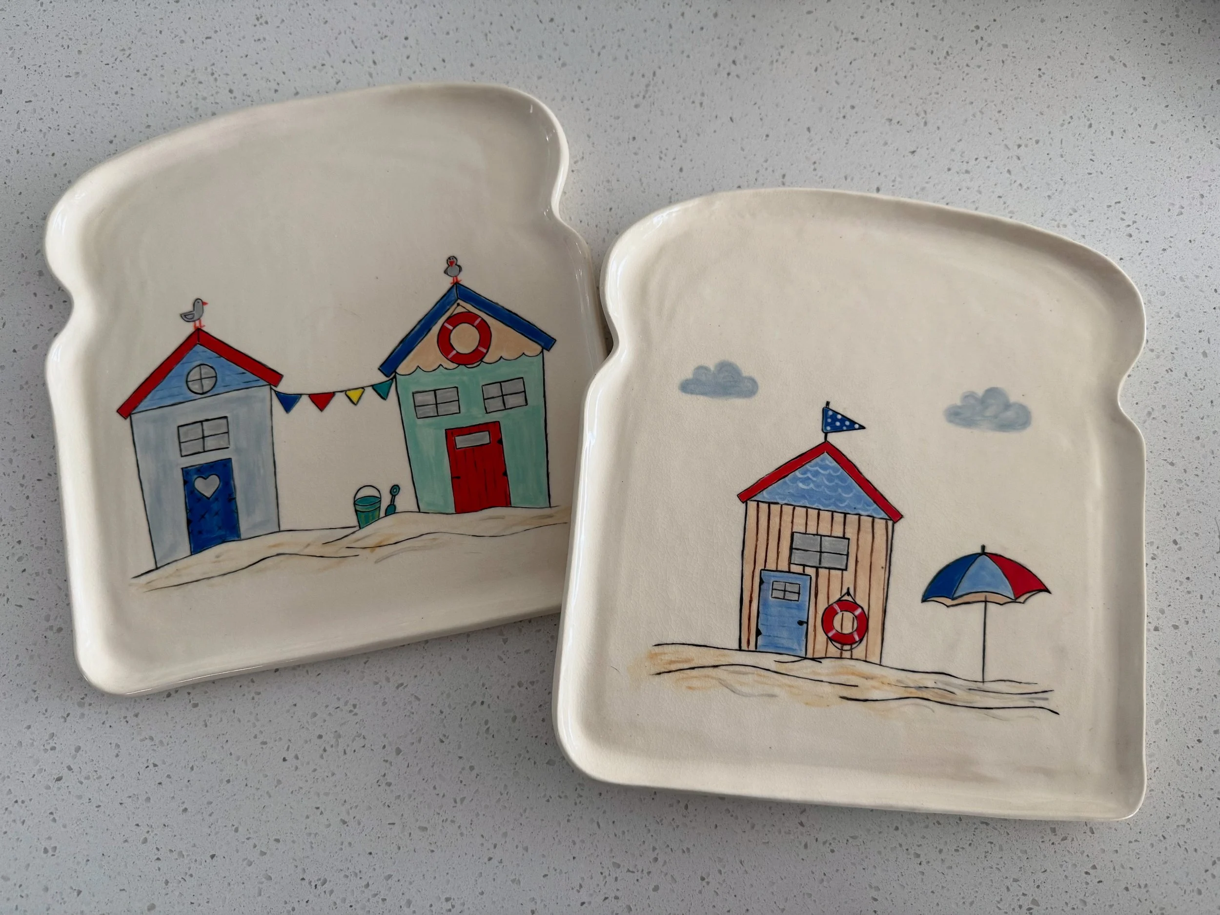 Two rectangular ceramic plates with rounded edges, decorated with seaside-themed illustrations. The left tray depicts two beach huts with flags, a basket, and a seagull. The right tray shows one beach hut with an umbrella and clouds in the sky.