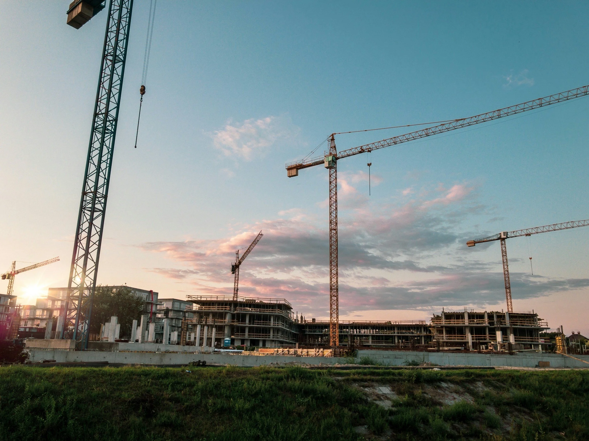 Construction site with multiple cranes and partially built structures under a partly cloudy sky during sunset.