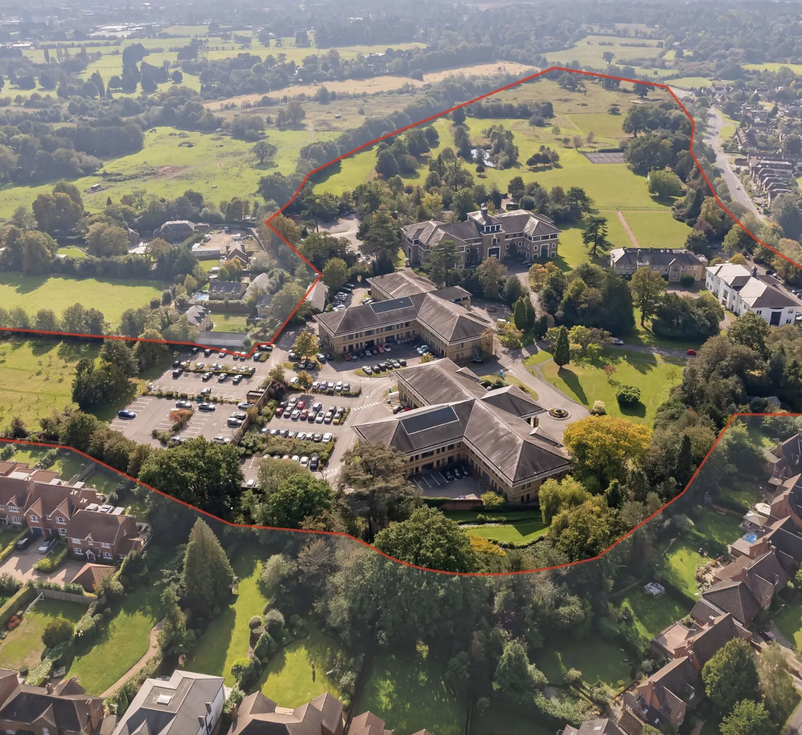 Aerial view of a large estate with multiple buildings, parking lots, and vast green lawns, surrounded by trees and residential neighborhood.