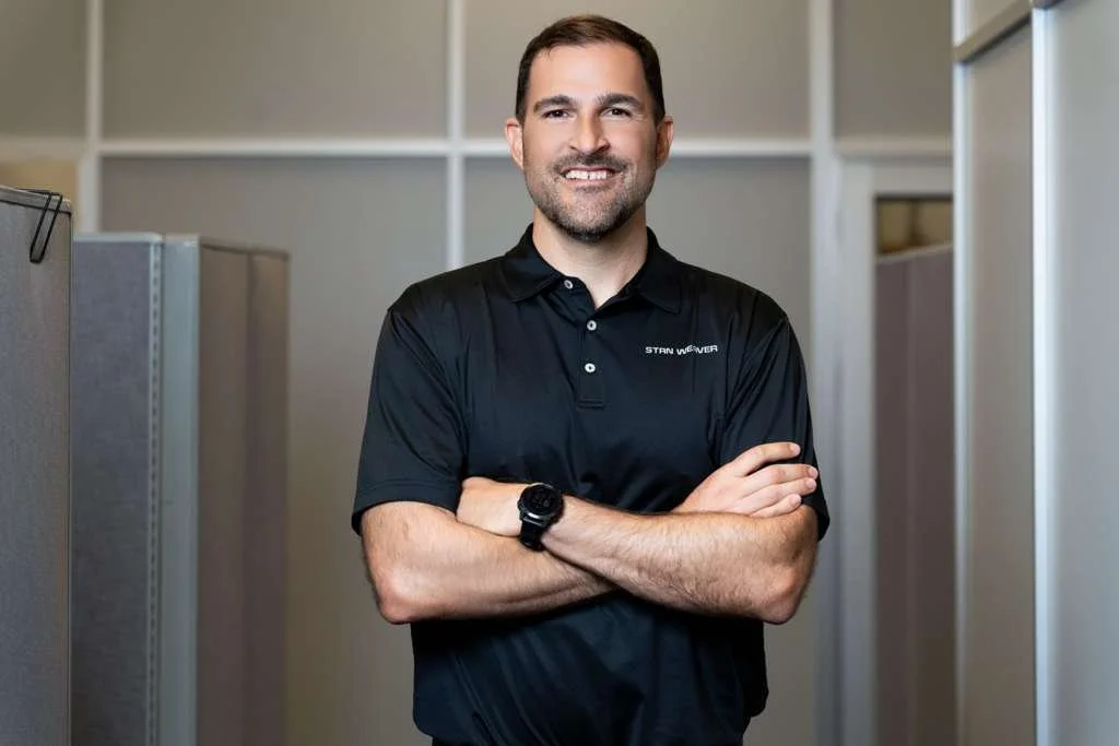 A man standing with arms crossed in an office cubicle area, wearing a black polo shirt and a watch, smiling at the camera.