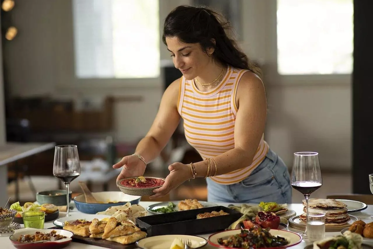A woman with dark hair, wearing a yellow and white striped tank top and light blue jeans, standing at a table with various dishes and red wine glasses. She is placing a dish on the table, smiling slightly.