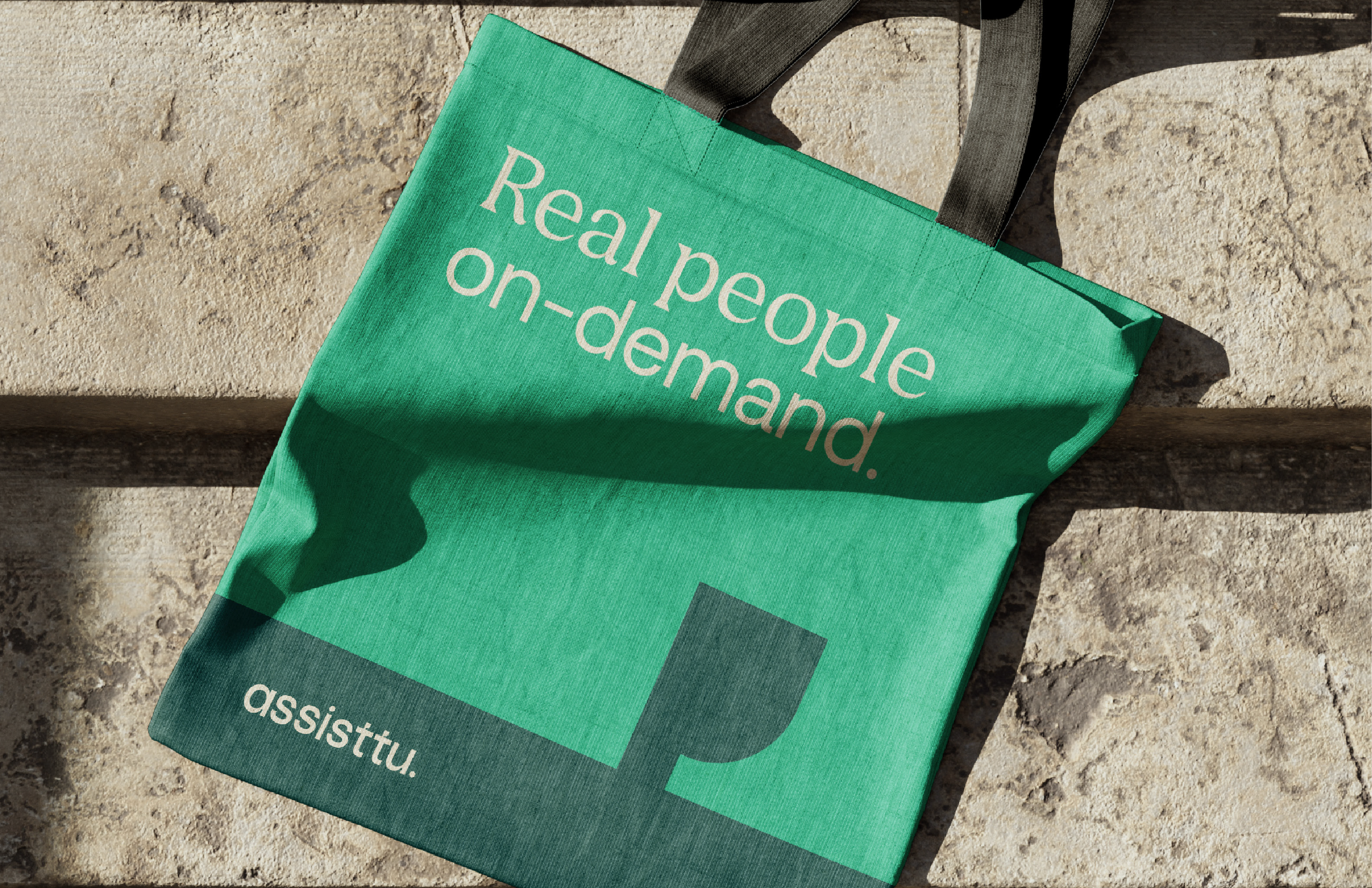 A green tote bag with the text 'Real people on-demand.' and the word 'assistu.' on it, lying on a stone surface.