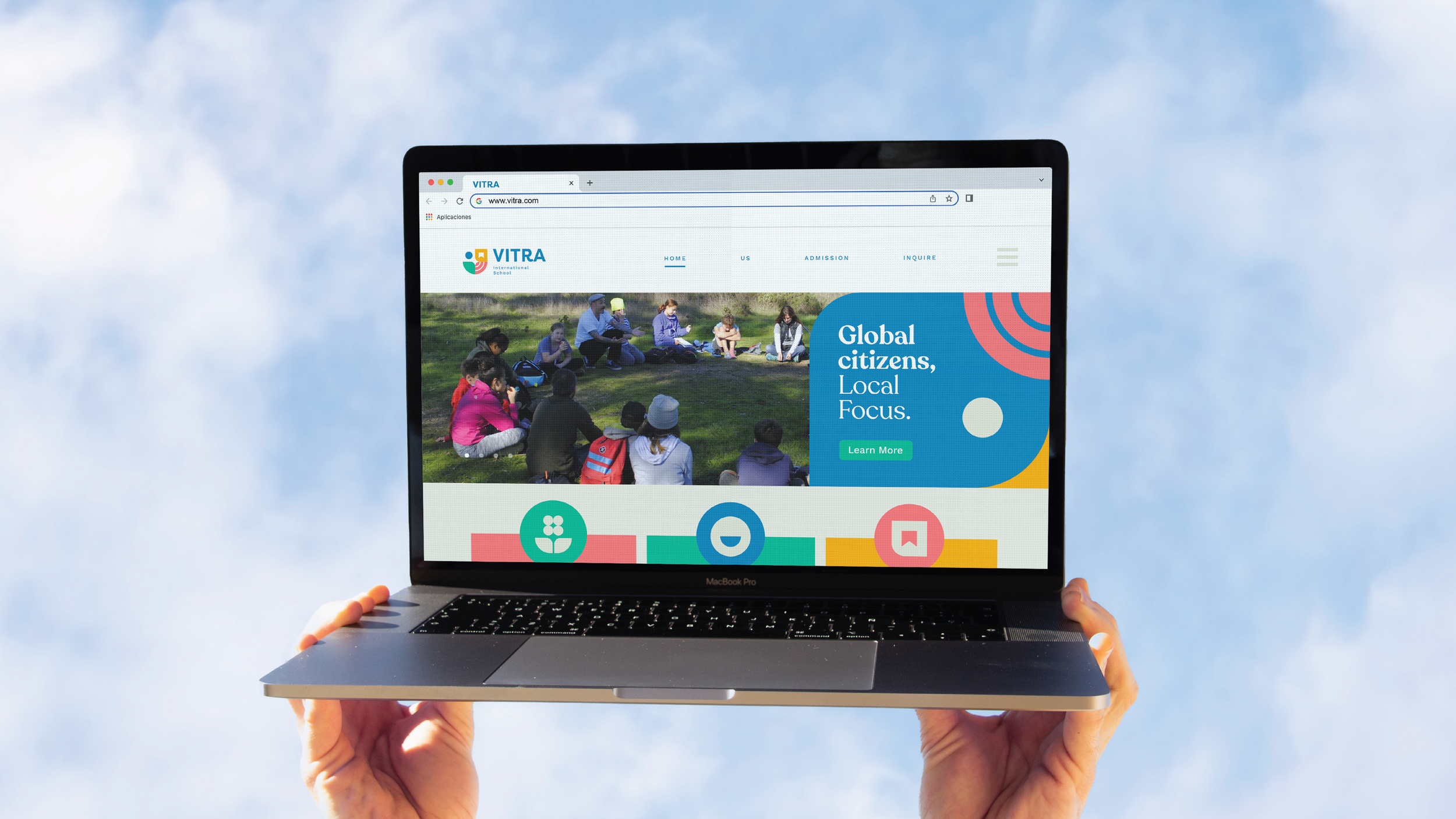 Person holding a MacBook Pro displaying a website titled 'VITRA' with a photo of children and adults sitting outdoors and text 'Global citizens, Local Focus' on a blue background.