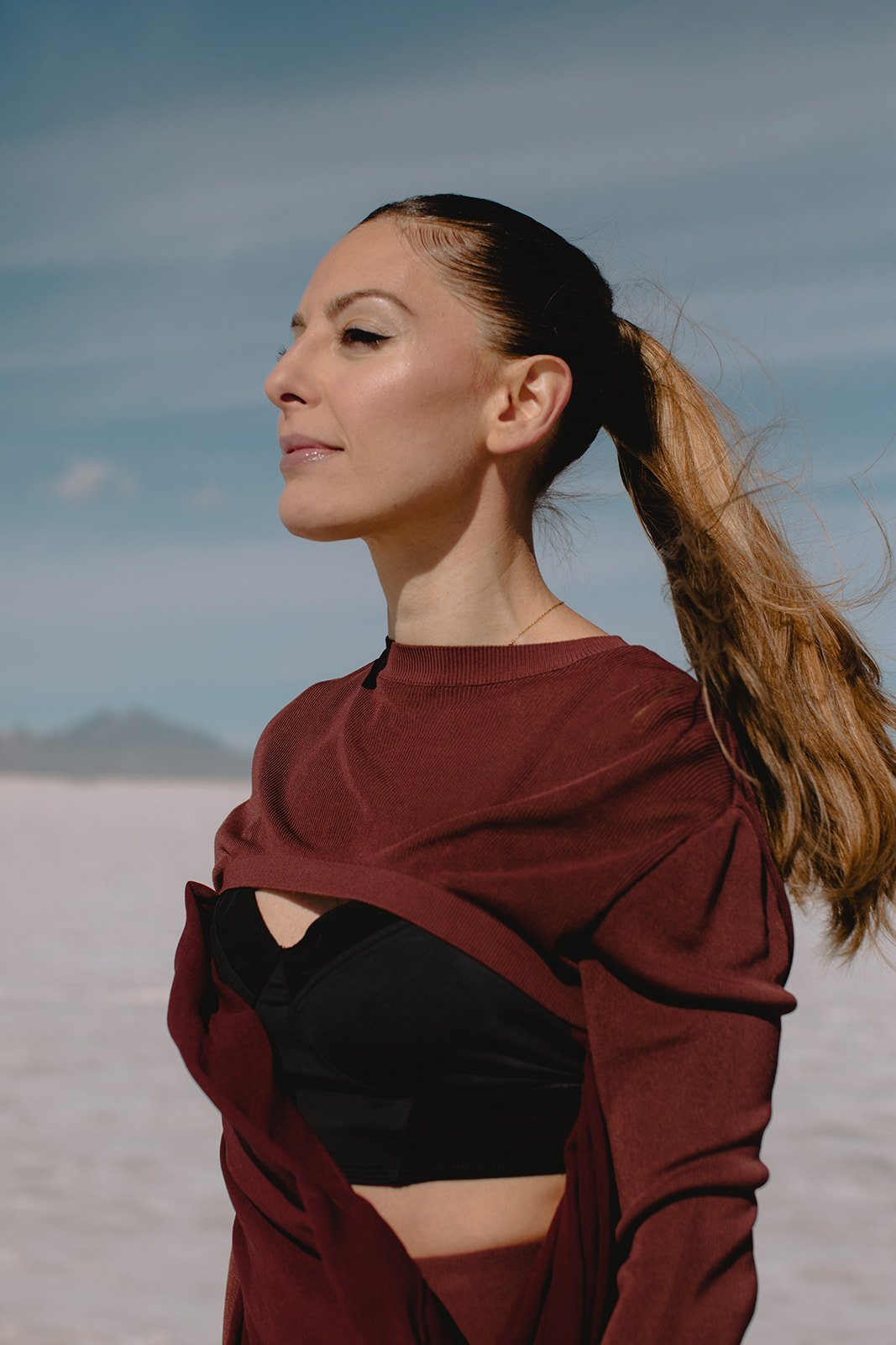 Paula Celestino with closed eyes and a relaxed expression, stands outdoors on a sandy background under a blue sky, wearing a maroon shirt over a black sports bra, with her hair tied back.