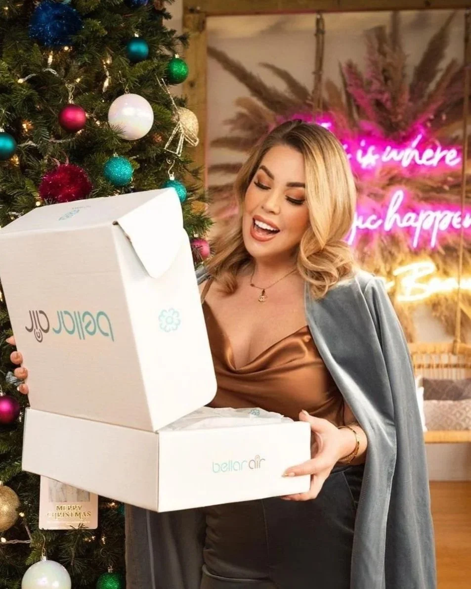 A happy influencer woman opening a white box labeled Bellair Air next to a decorated Christmas tree with ornaments. She appears excited as she takes out a product from the box indoors, with a pink neon sign in the background.