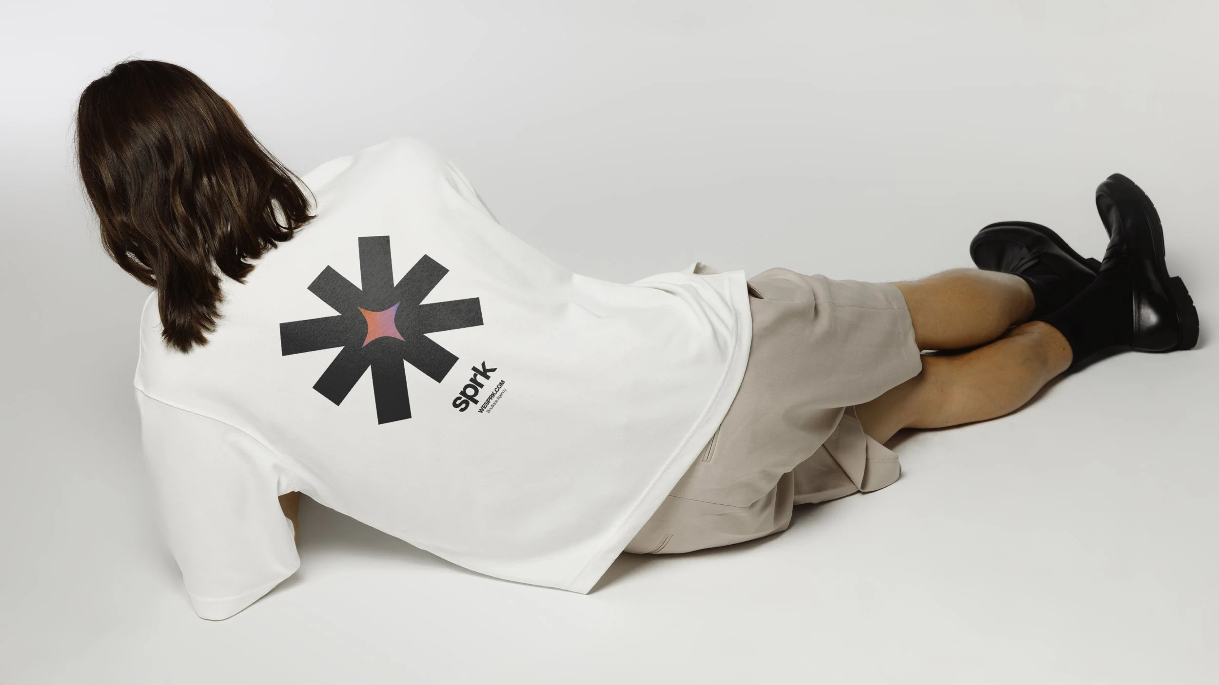 Person lying on the floor with a white t-shirt and black shoes, wearing beige shorts, facing away, against a plain white background.