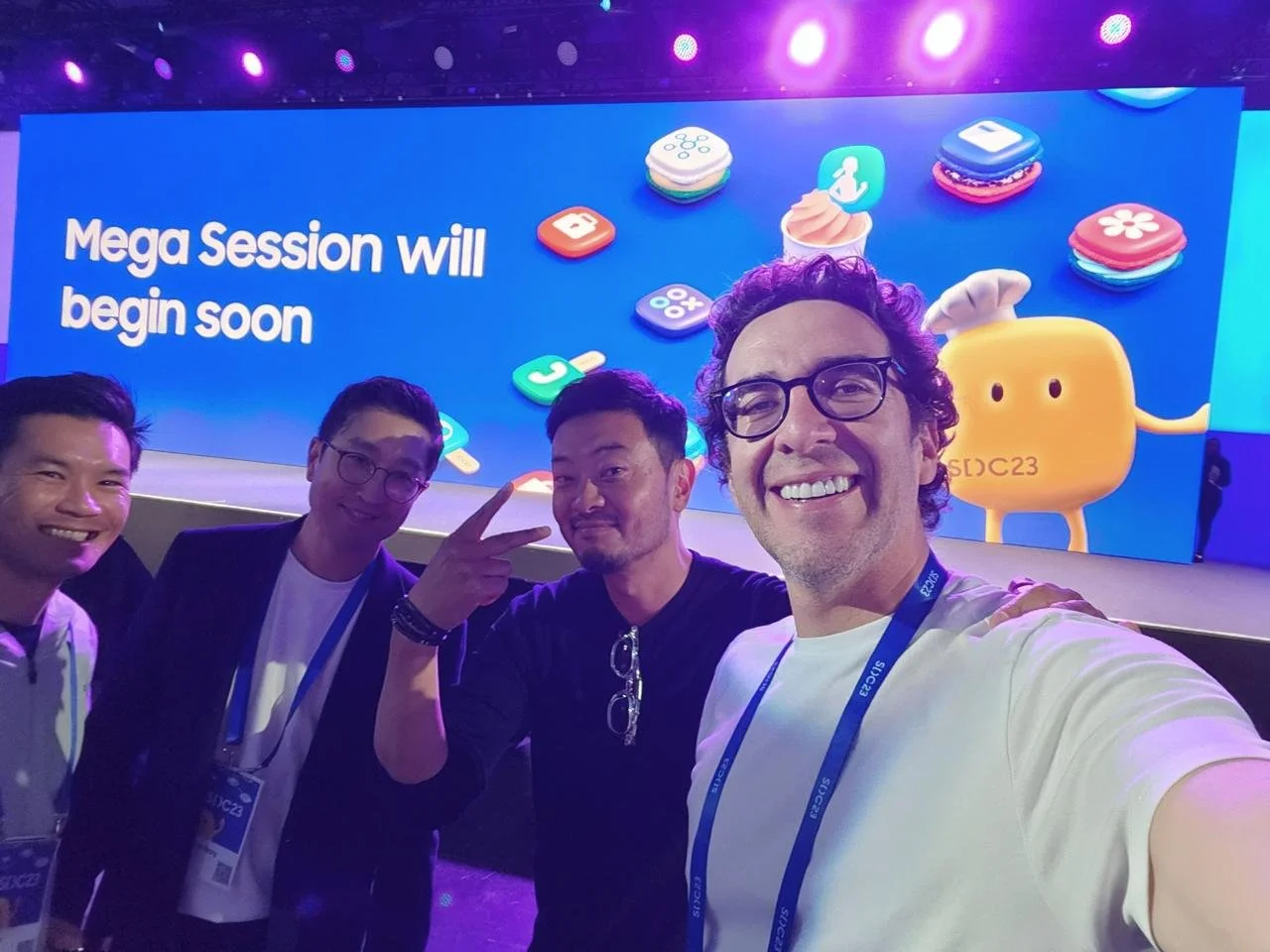 Four men taking a selfie at a conference stage with a large screen behind them displaying the text "Mega Session will begin soon" and colorful app icons. The men are smiling, and one is making a peace sign.
