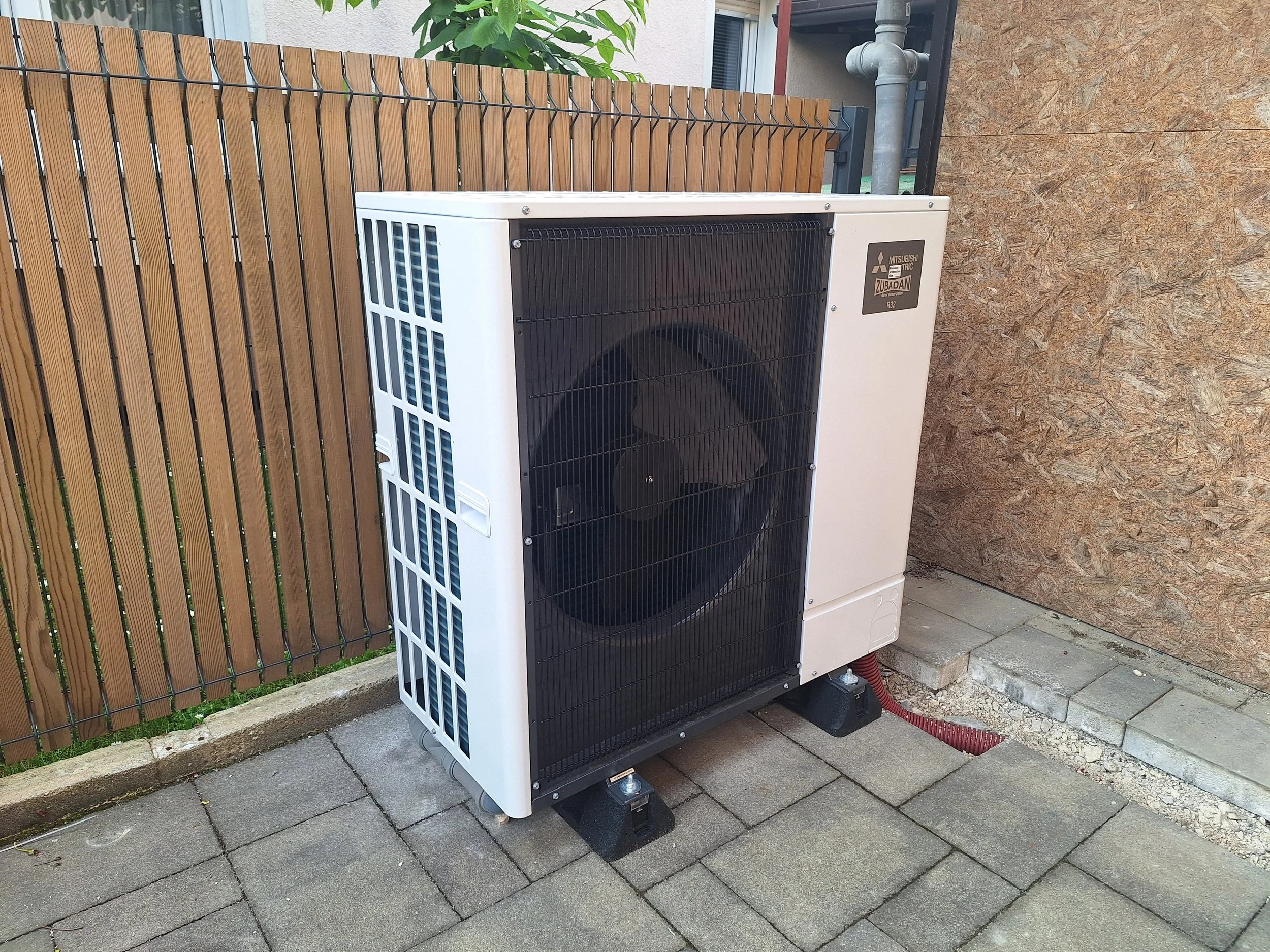 Zunanja klimatska naprava Mitsubishi z zaščitno rešetko, postavljena na betonski plošči blizu temno lesene stene, z majhno vedro in orodjem v bližini.