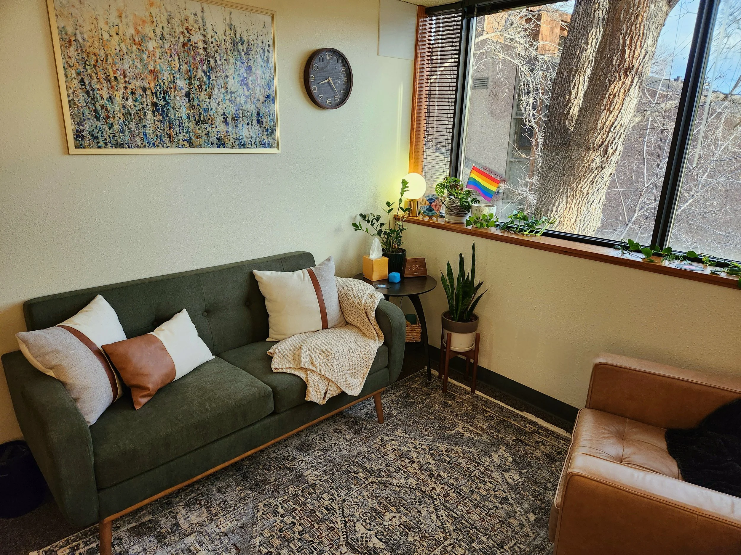 Living room with a green sofa, a tan armchair, a patterned rug, a windowsill with plants, a rainbow pride flag, a small side table with a lamp, and a large abstract painting on the wall.