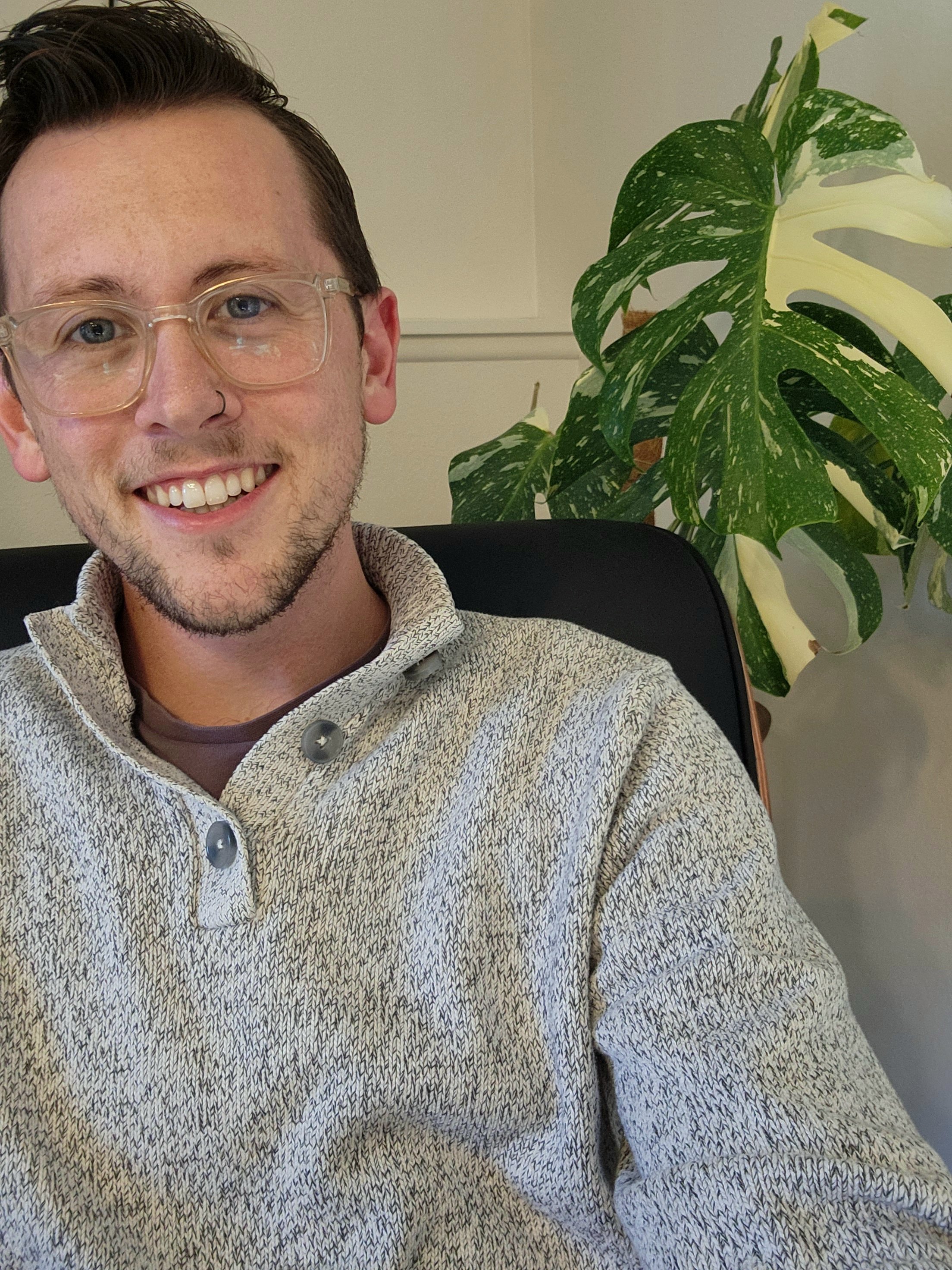 A young man with glasses and a nose ring smiling, sitting in a black office chair, with a large green and white variegated monstera plant in the background.