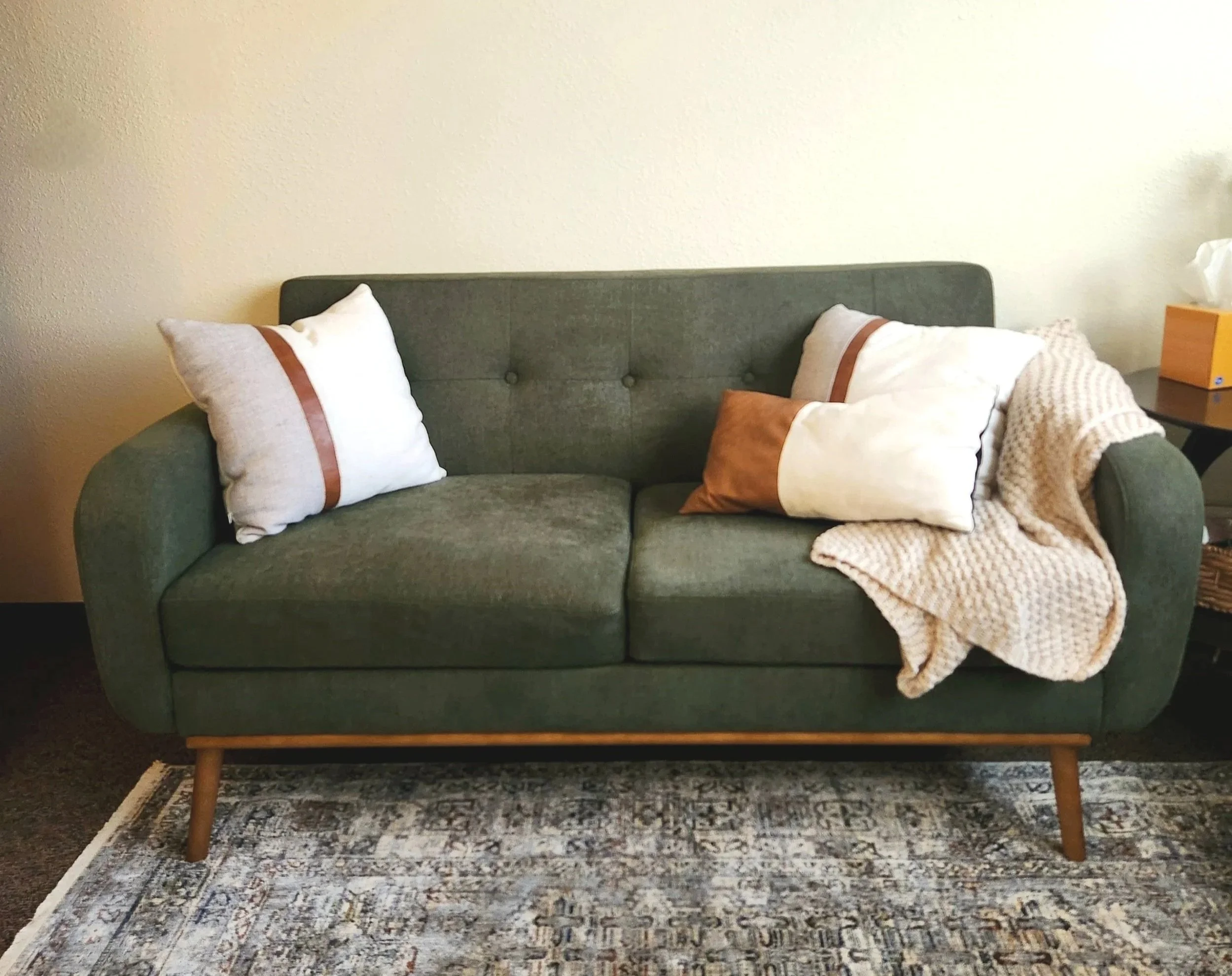 Warm and inviting counseling office featuring a green mid-century modern sofa, abstract artwork, textured rug, and neutral accent pillows for a comfortable therapy setting.