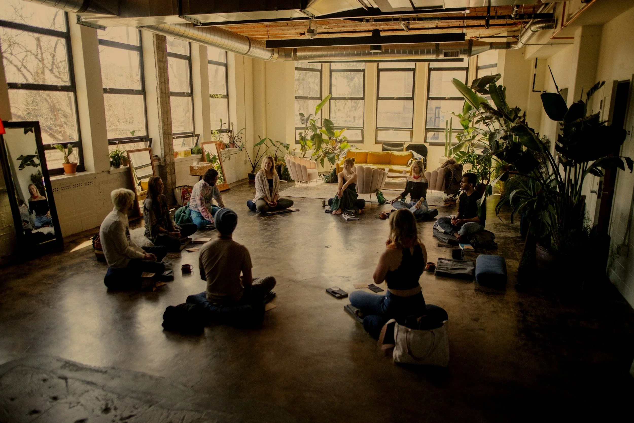People sitting in a circle on the floor in a well-lit room with large windows, plants, and minimal decor, participating in a group activity or meditation session.