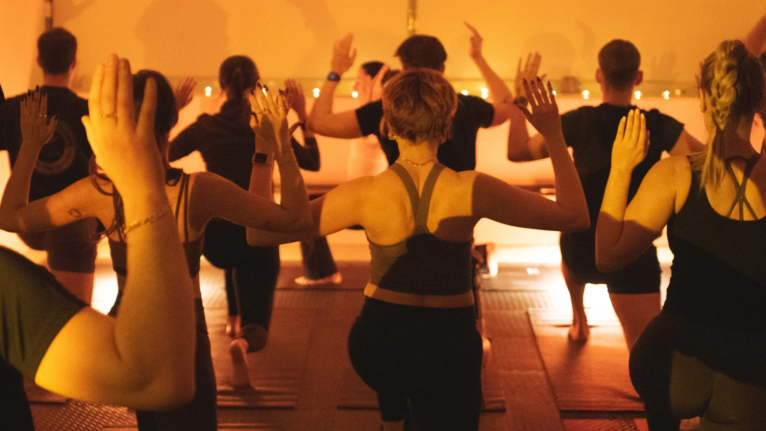People sitting in a circle on the floor in a well-lit room with large windows, plants, and minimal decor, participating in a group activity or meditation session.