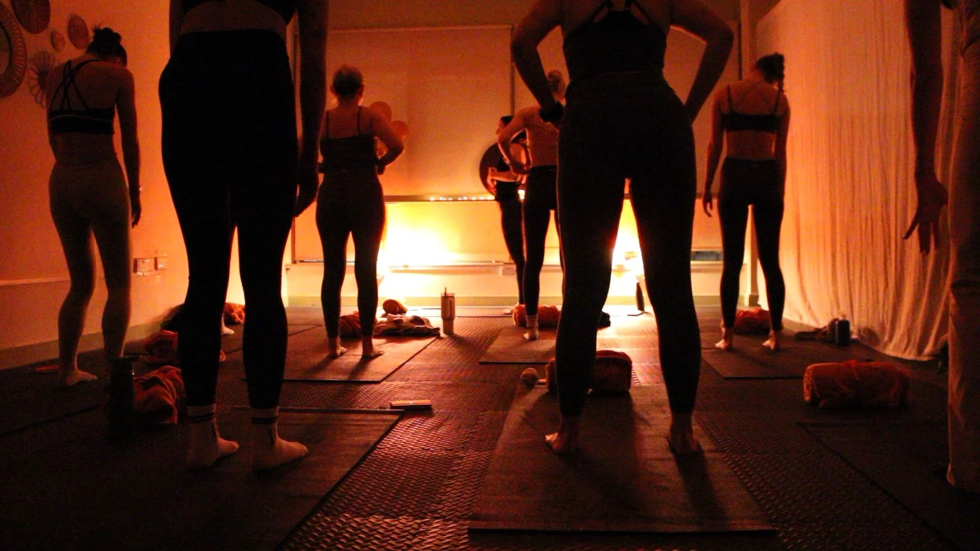 Group of people in a dimly lit room participating in a yoga or meditation class, facing a warm light source at the front, with mats and water bottles on the floor.