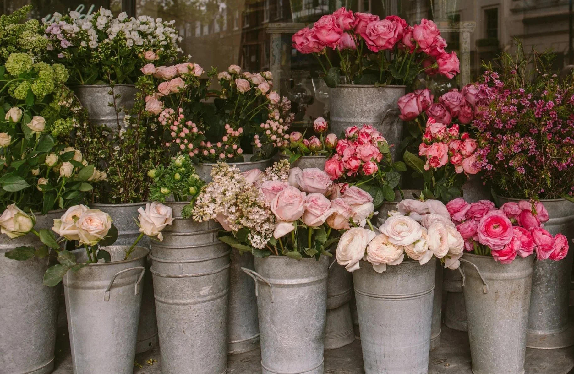 A display of various pink and white flowers in metal buckets outside a shop window.