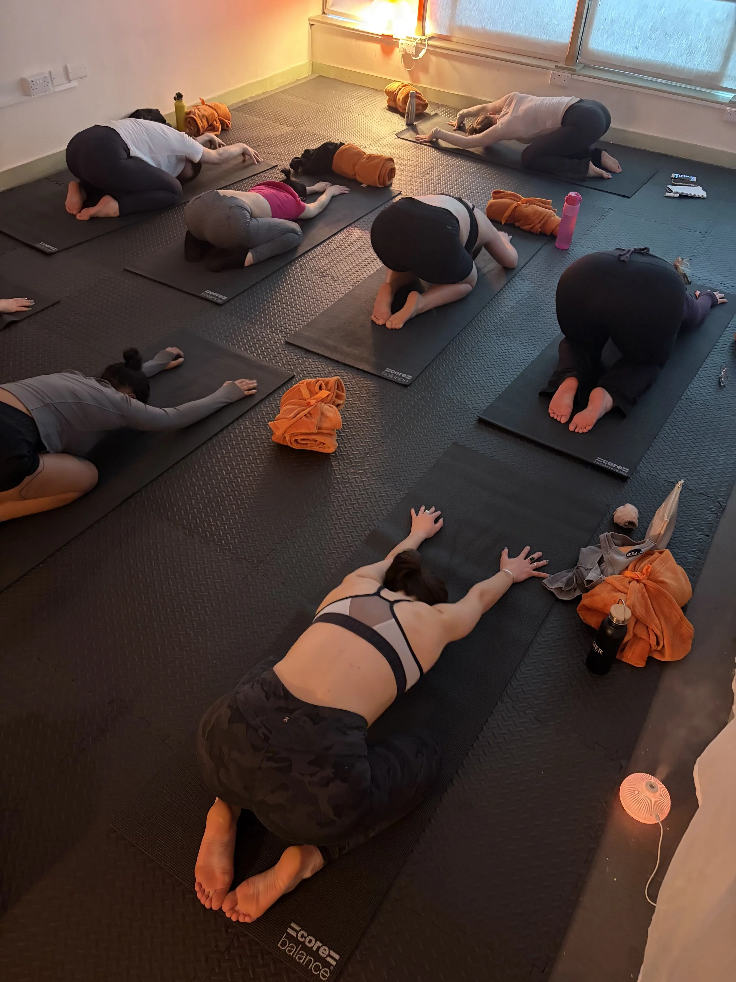 Group of people participating in a yoga or meditation class on black mats in a room with soft lighting and large windows.