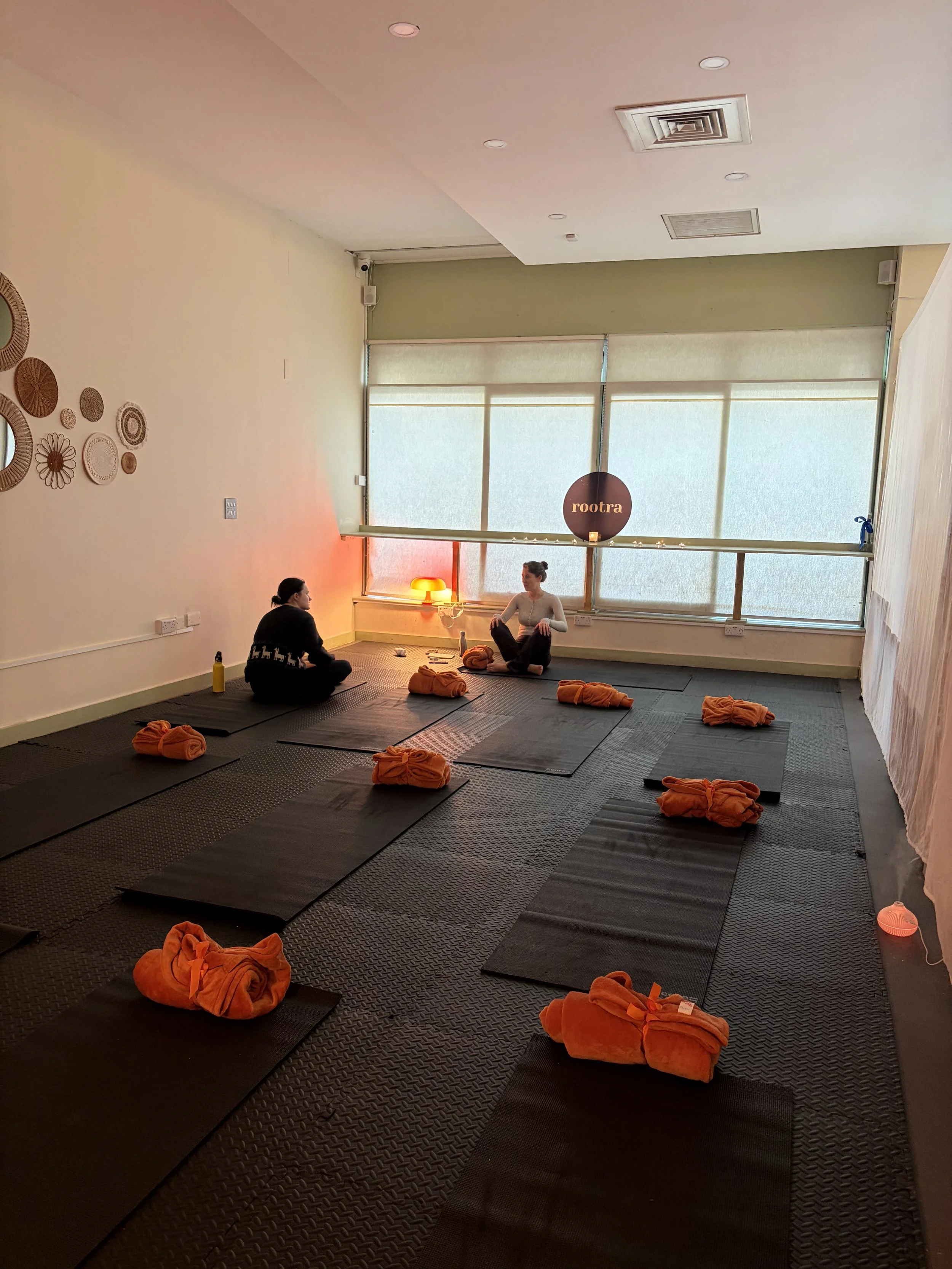 Indoor yoga studio with black mats and orange towels folded on each, two women sitting on mats, one facing the camera and one with her back to it, a window with blinds, a sign that reads "rootra" in the background, and decorative wall art.