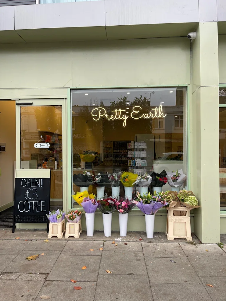 The storefront of Pretty Earth, a flower shop, displays various bouquets in white vases outside. A black chalkboard sign advertises open hours and coffee for £3. The shop has a large glass window and a sign with the business name in neon cursive.