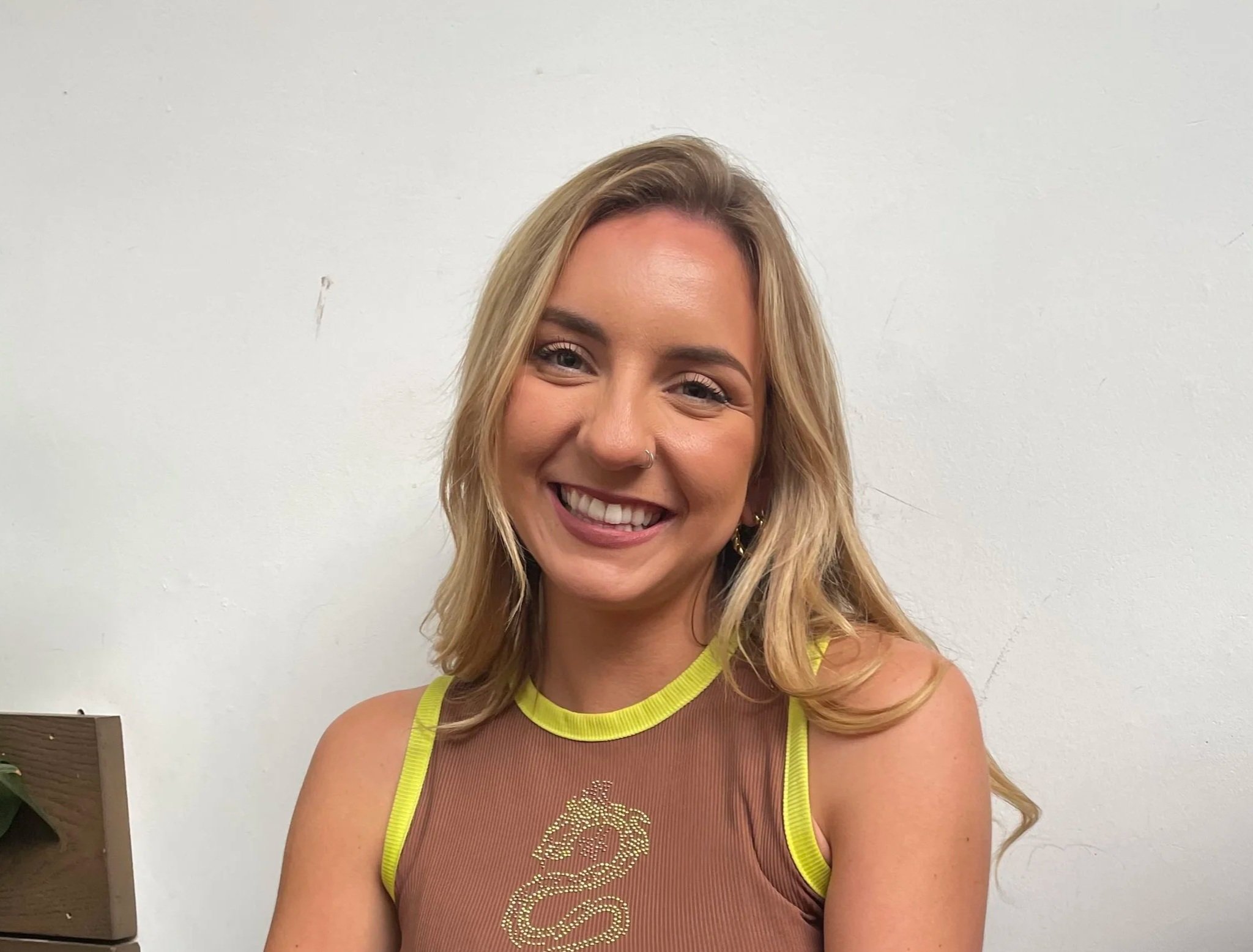 A young woman with shoulder-length blonde hair smiling, wearing a sleeveless brown top with yellow trim and a gold dragon design, standing against a white wall.