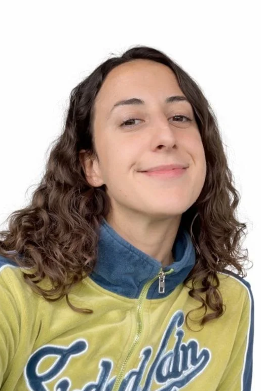 A woman with curly brown hair, wearing a yellow jacket with a blue collar, smiling slightly at the camera against a plain white background.