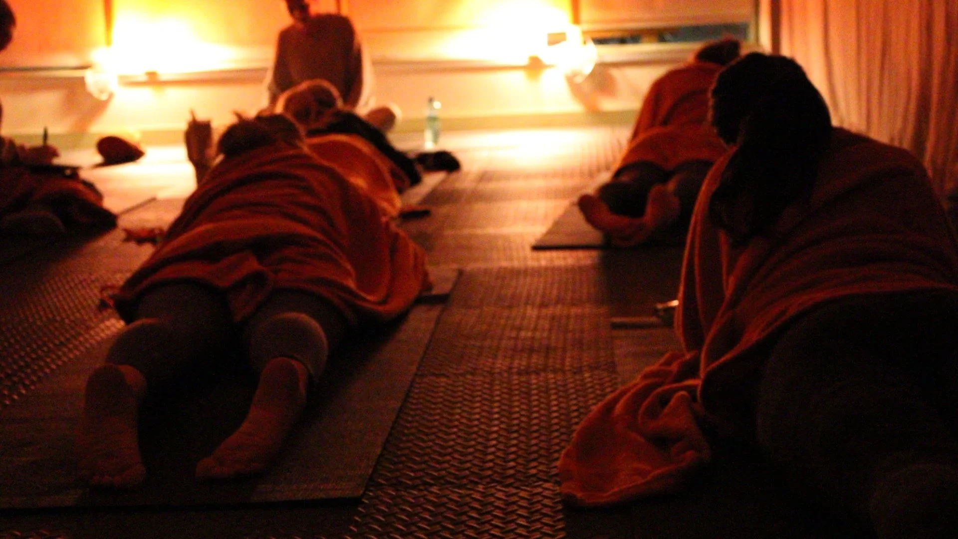 People lying on their stomachs in a dimly lit room, possibly practicing meditation or relaxation exercises, with a person standing at the front near a blackboard.