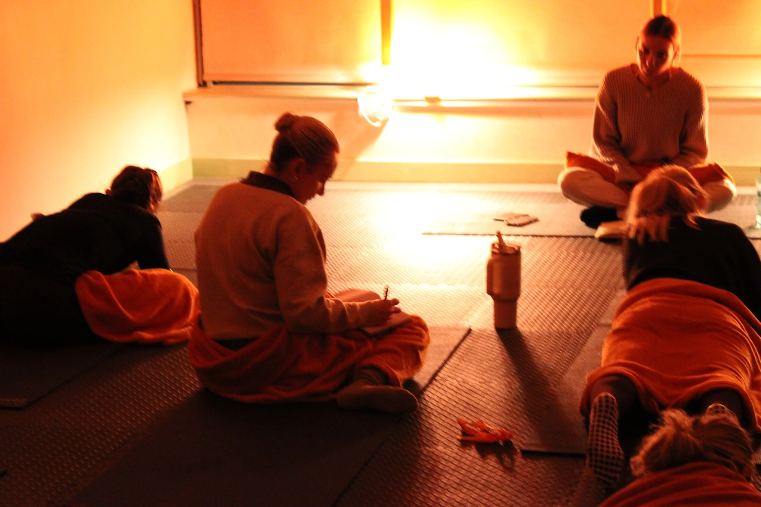 A group of people, including Buddhist monks and a woman, are sitting and lying on their knees on mats in a dimly lit room, engaging in meditation or prayer, with a small lit lamp in the background.