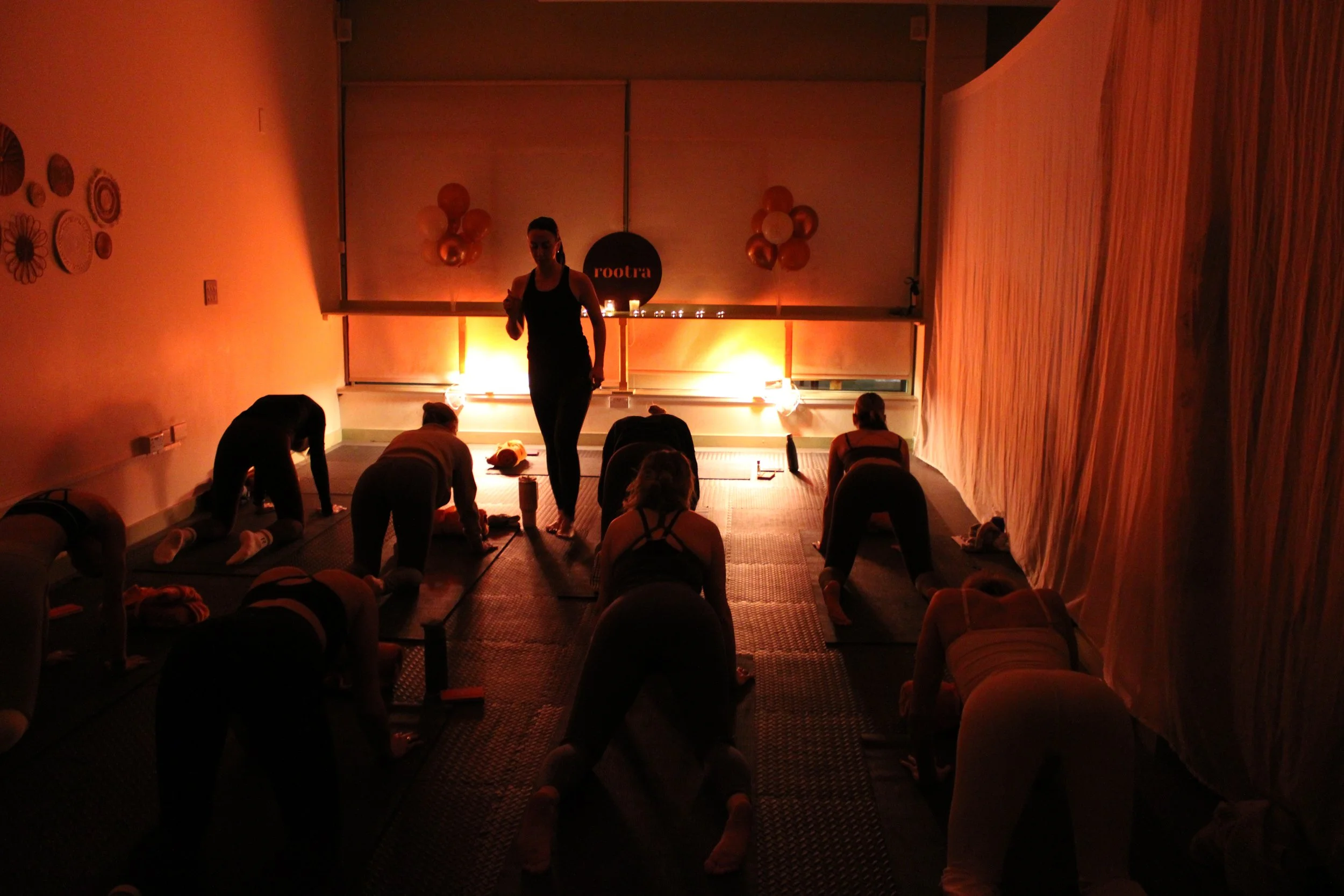 A group of people participating in a yoga class in a dimly lit studio, with a woman instructor in front and attendees on yoga mats, illuminated by warm orange lighting.