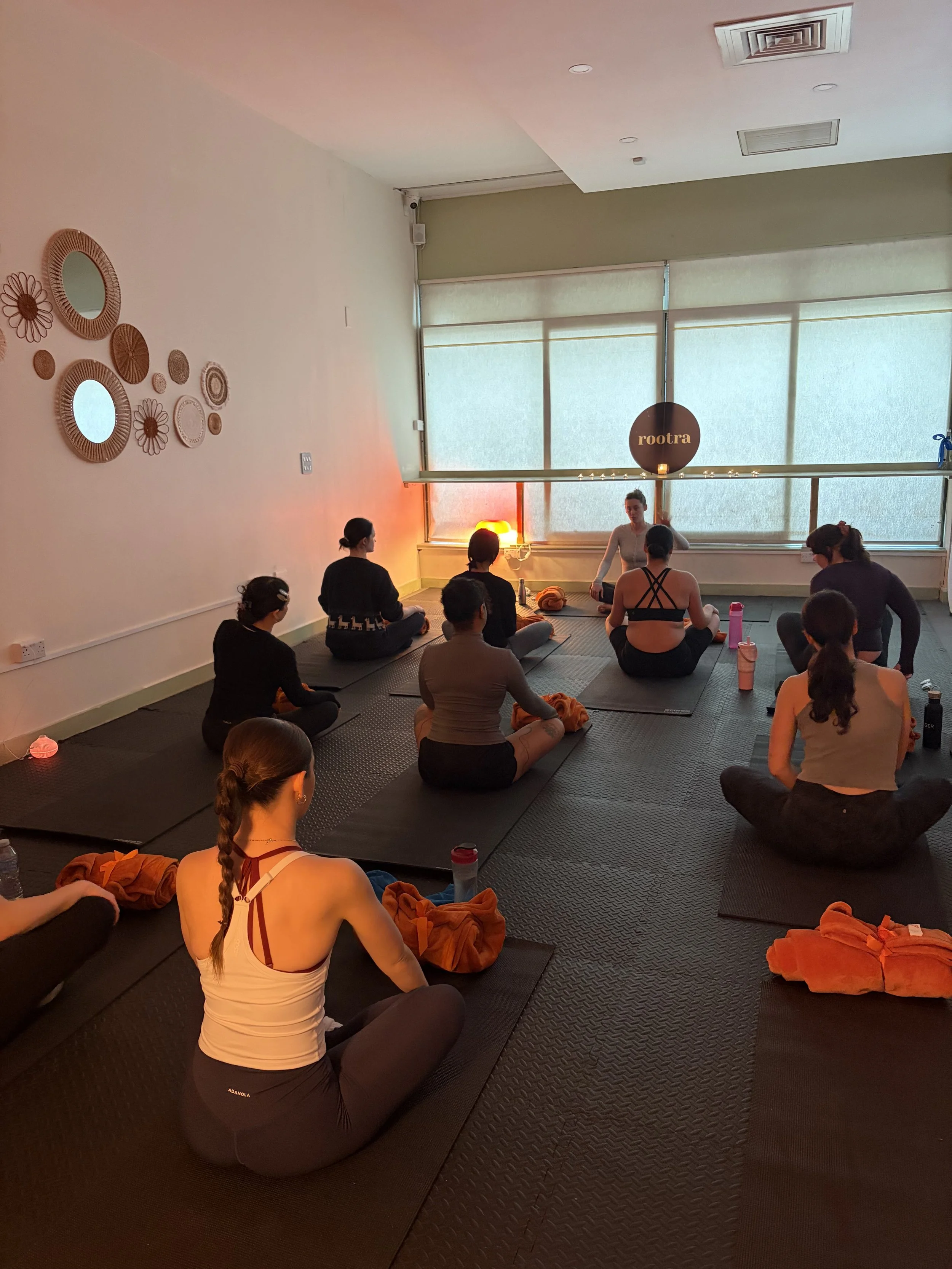 A group of people participating in a yoga class in a studio with large windows, black mats, and decorations on the wall.