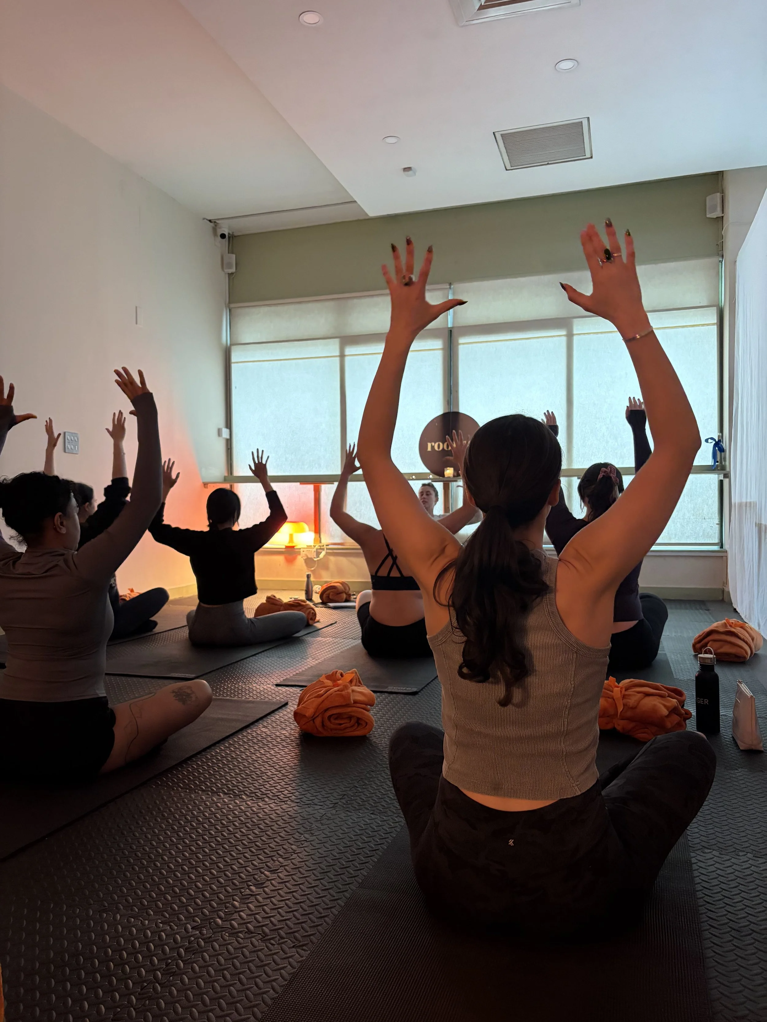 Group of people participating in a yoga or meditation class in a calm, softly lit room, sitting on mats with their hands raised in a gesture of relaxation or stretching, with large windows in the background.