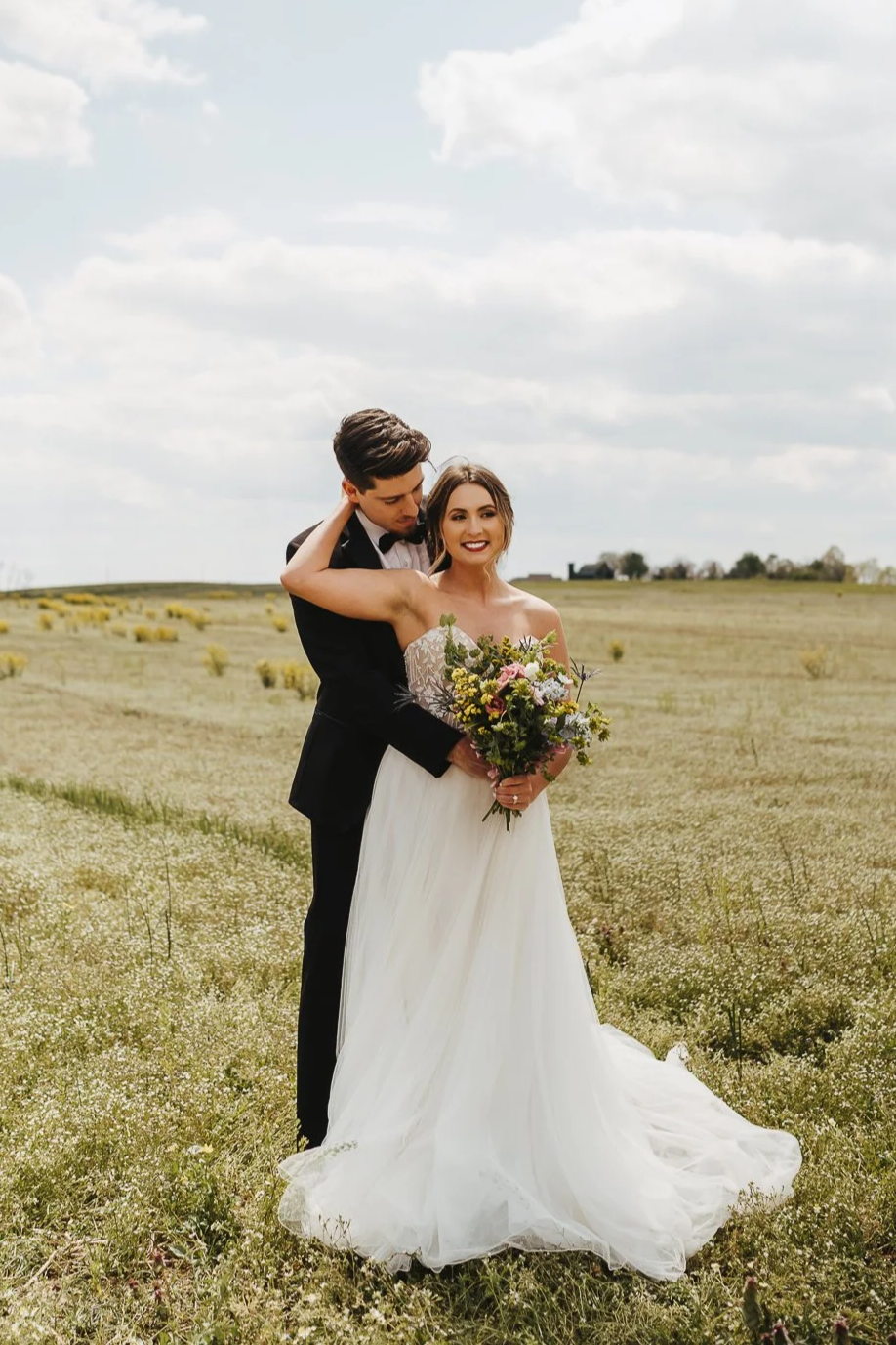 A smiling bride holding a bouquet of flowers, standing in a grassy field with a groom in a black suit | Videography and Photography | Evansville, IN