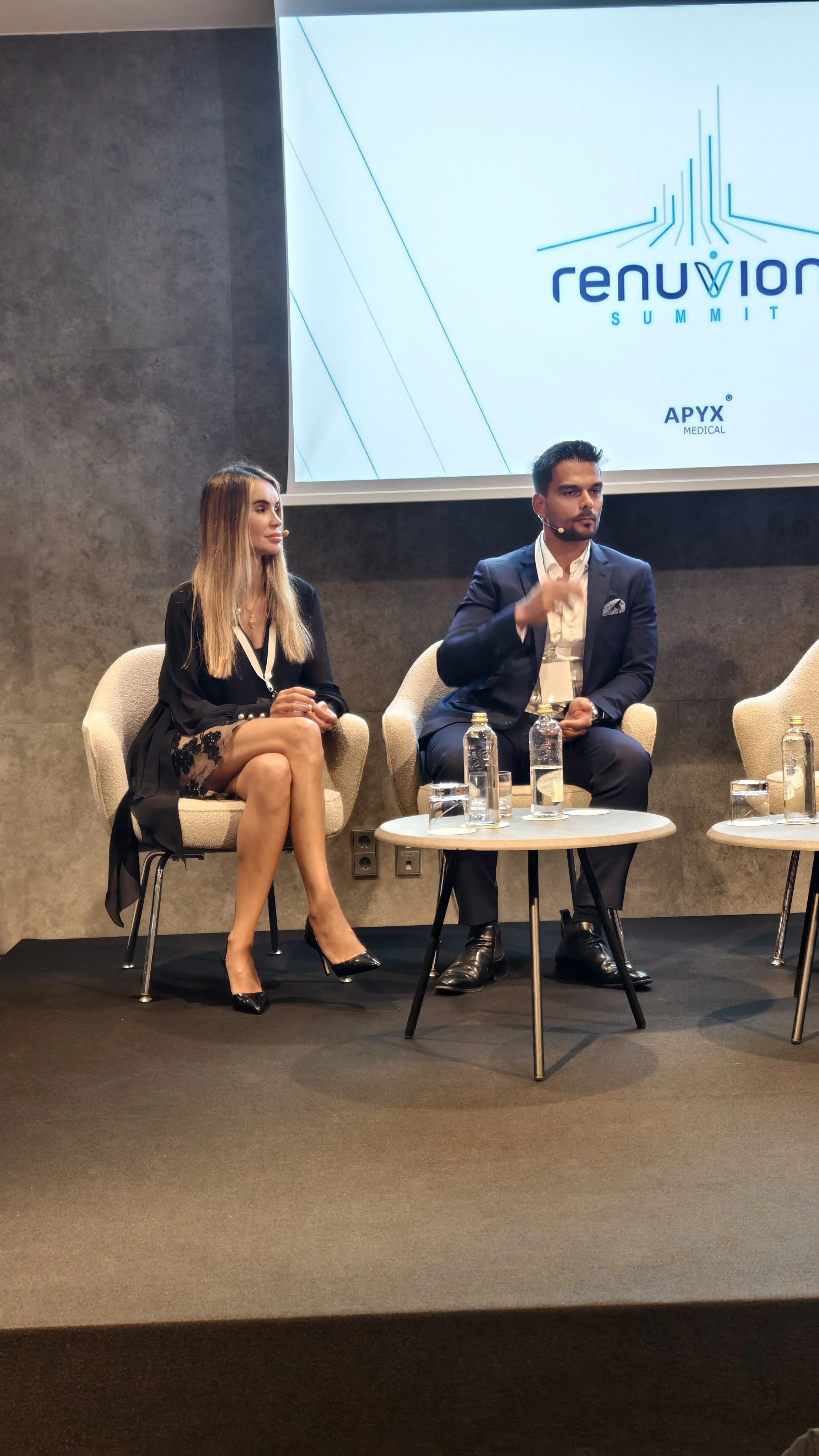 Dois pessoas em uma mesa de painel, uma mulher e um homem, participando de um evento. A mulher veste vestido preto com salto alto, e o homem usa terno escuro. Ambos usam microfones de headset. No fundo, tela com logo do evento "Renuvion Summit".
