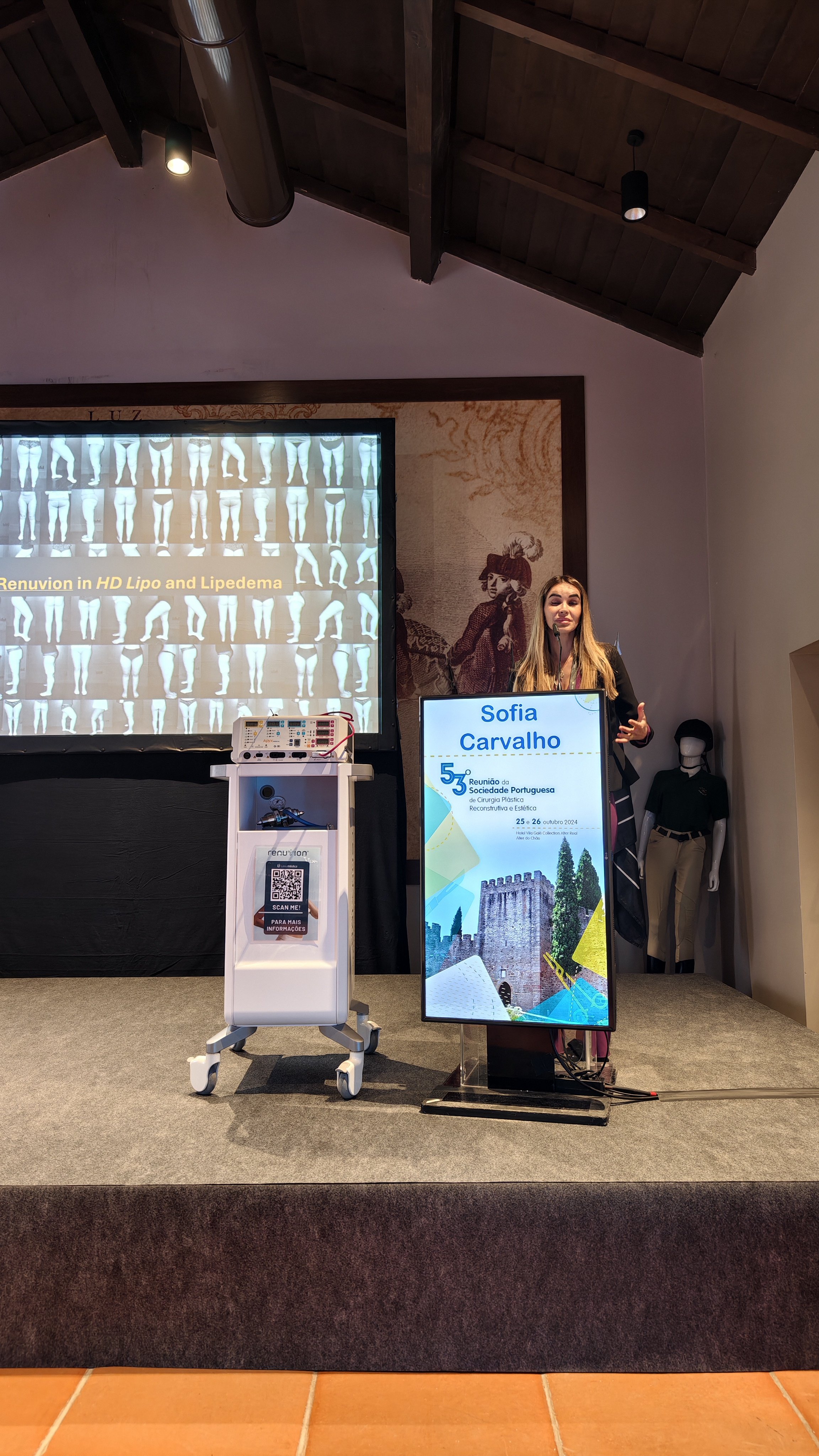Mulher apresentando em palco durante uma conferência, com uma tela ao fundo mostrando imagens de exames de radiografia e um painel digital ao seu lado exibindo o nome Sofia Carvalho. Há uma manequim com roupas ao lado direito da apresentadora.