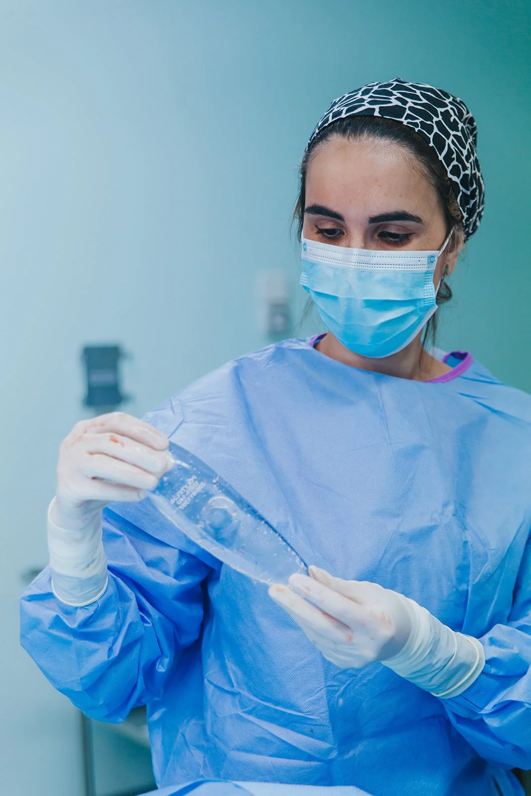 Profissional de saúde usando máscara facial, luvas e jaleco azul, manipulando uma ampola de material médico ou farmacêutico em ambiente hospitalar.