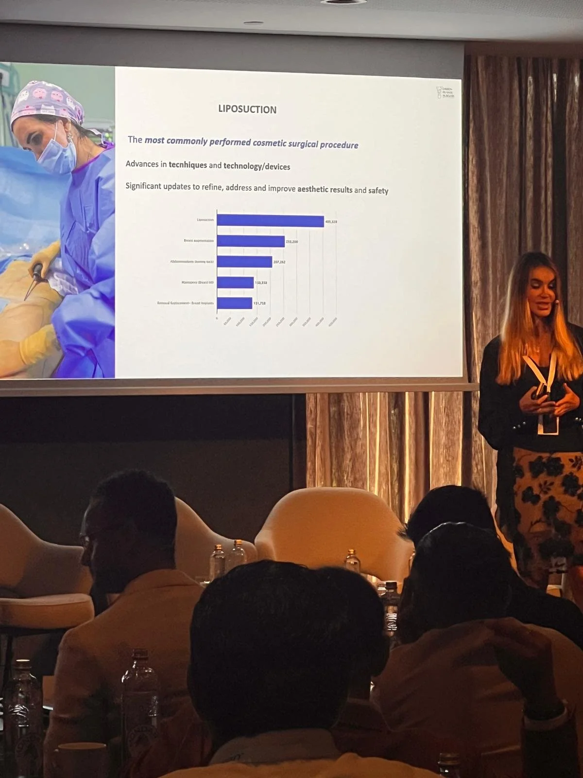 Slide de apresentação sobre lipoaspiração, com uma imagem de uma cirurgiã vestida com roupas cirúrgicas e máscara, ao lado de um gráfico de barras e texto em inglês. A apresentadora está diante de uma sala com pessoas assistindo.