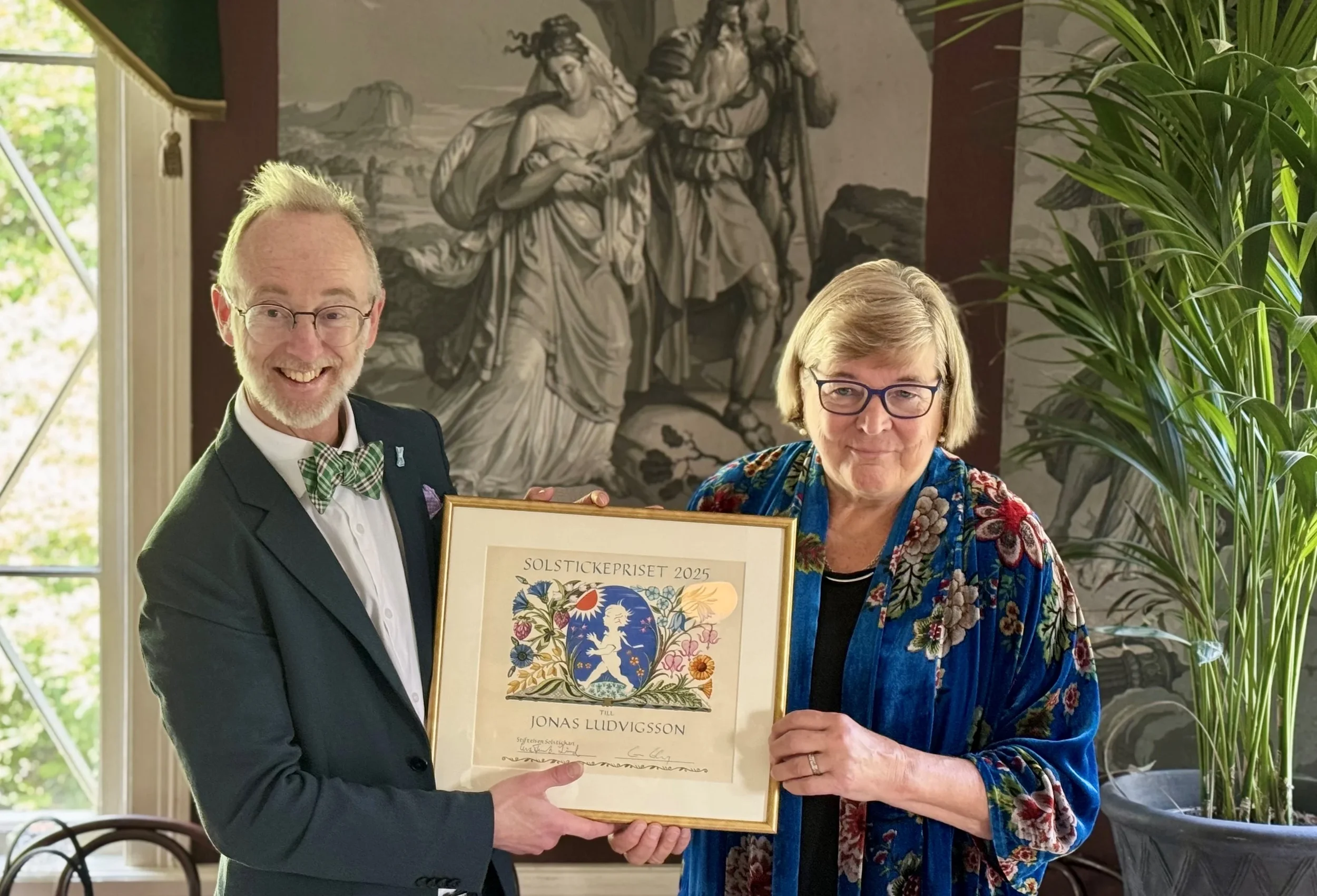 A man and woman holding a framed certificate together indoors, with a large black and white mural on the wall and a potted plant nearby, both smiling at the camera.
