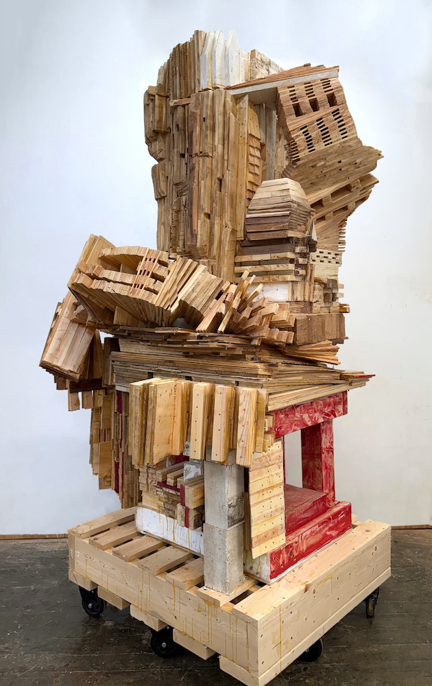 A large abstract sculpture made of various wooden pieces stacked and arranged on a wooden pallet on wheels, with no clear subject or recognizable form.