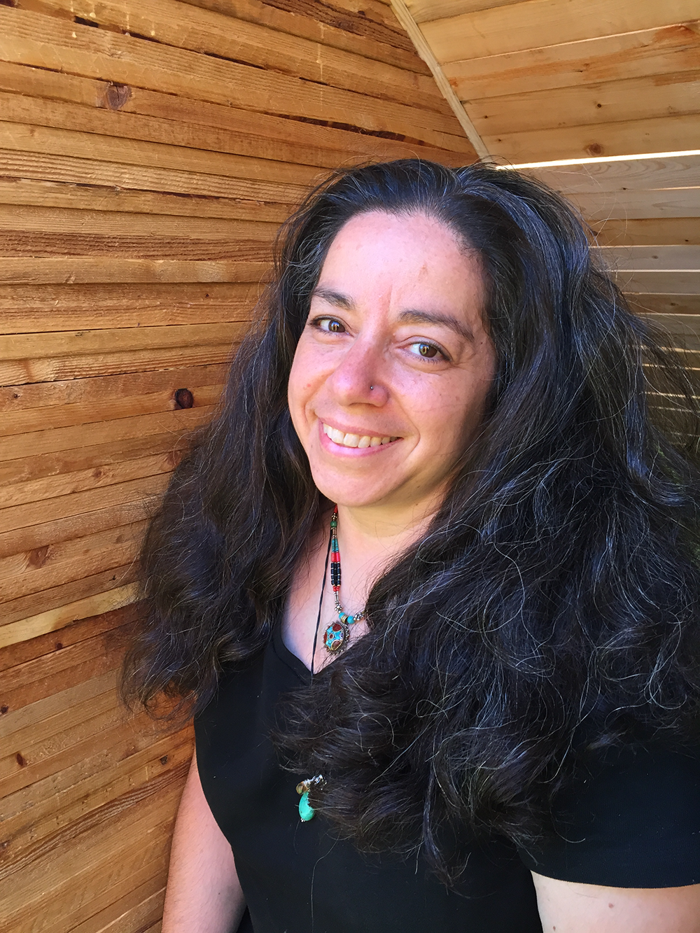 A woman with long, wavy black hair smiling, wearing a black top and colorful jewelry, against a wooden wall background.