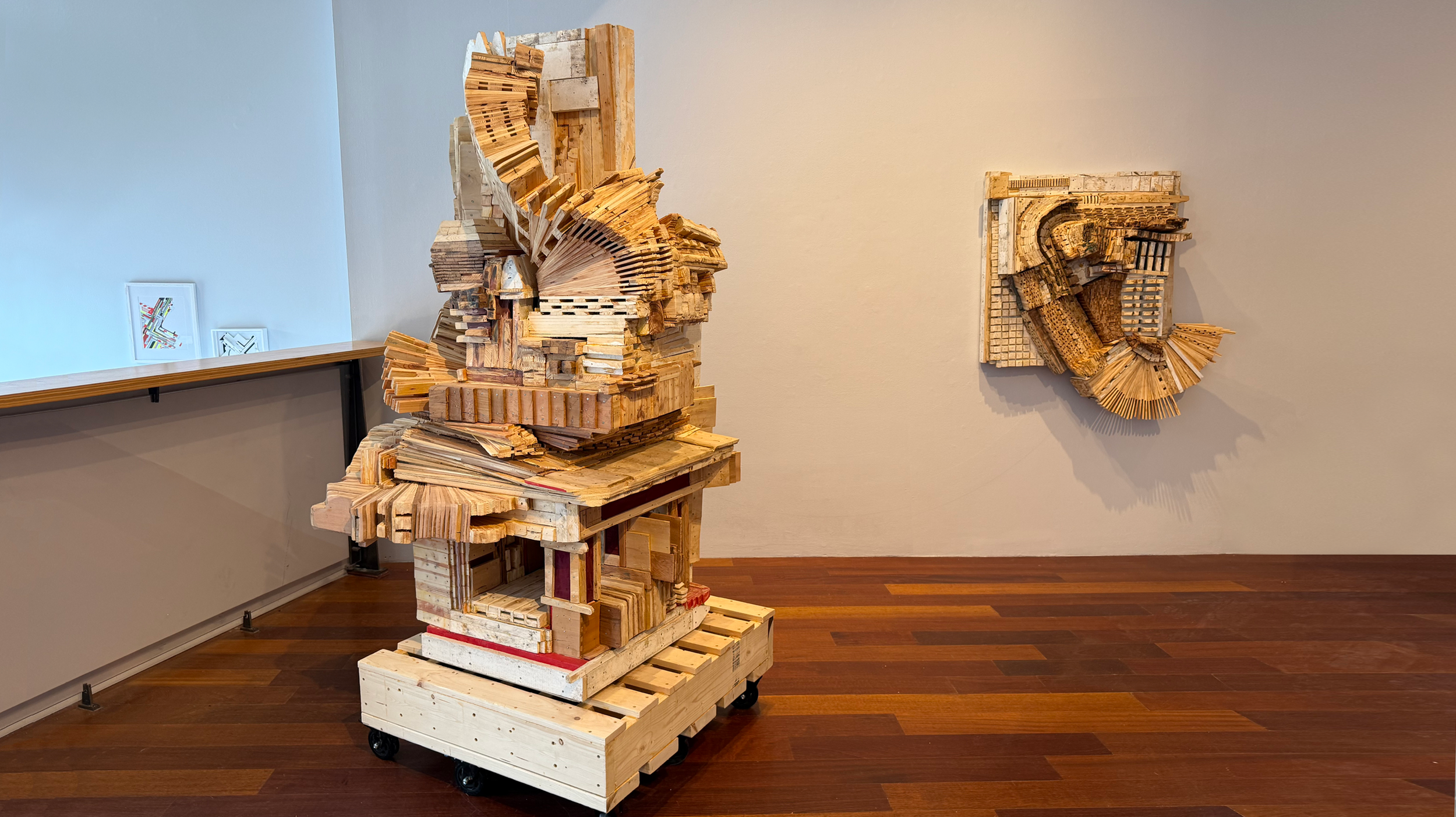 Contemporary art installation of abstract wooden sculptures in an art gallery, with one large multi-layered piece on a wheeled pallet and another smaller piece on the wall.