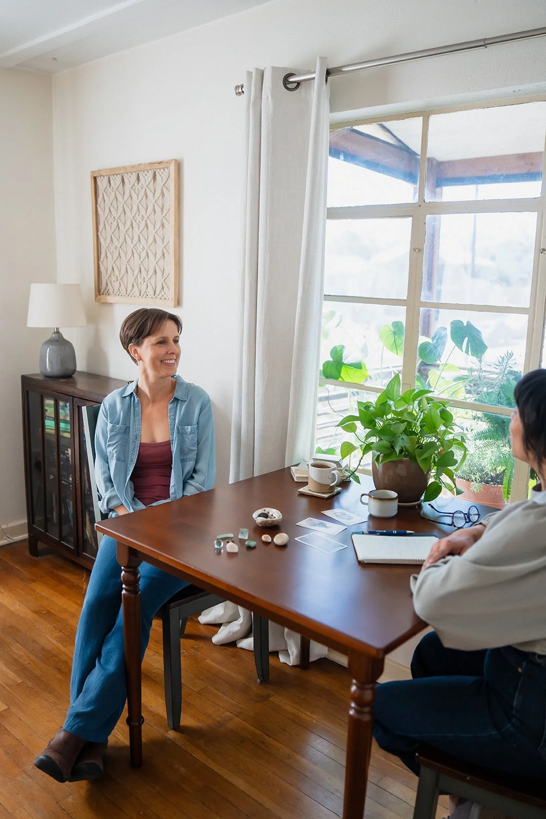 Two women having a conversation at a wooden dining table in a bright room with large window and green plants.