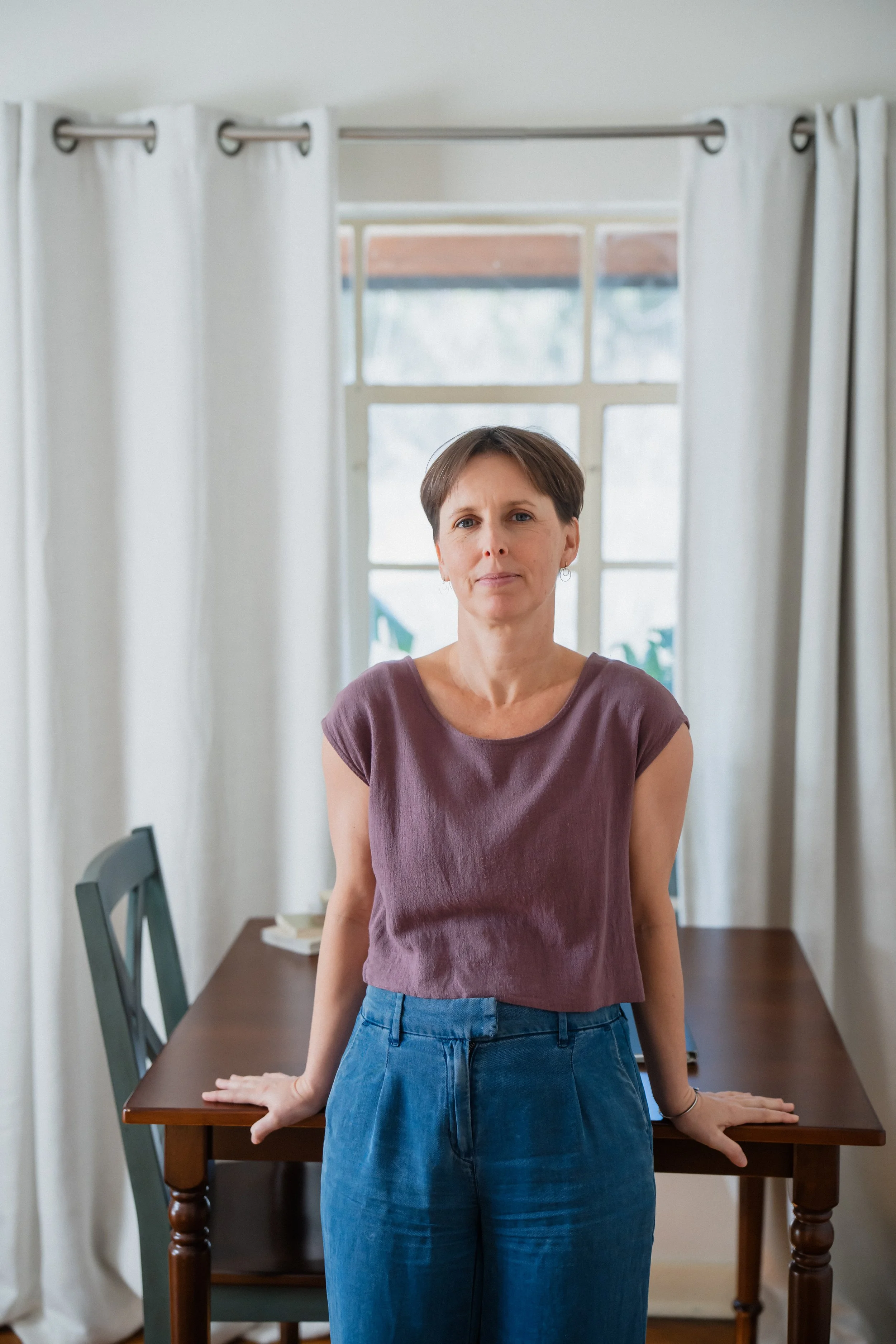 A woman standing behind a wooden table with her hands resting on it, in a room with white curtains and large windows.