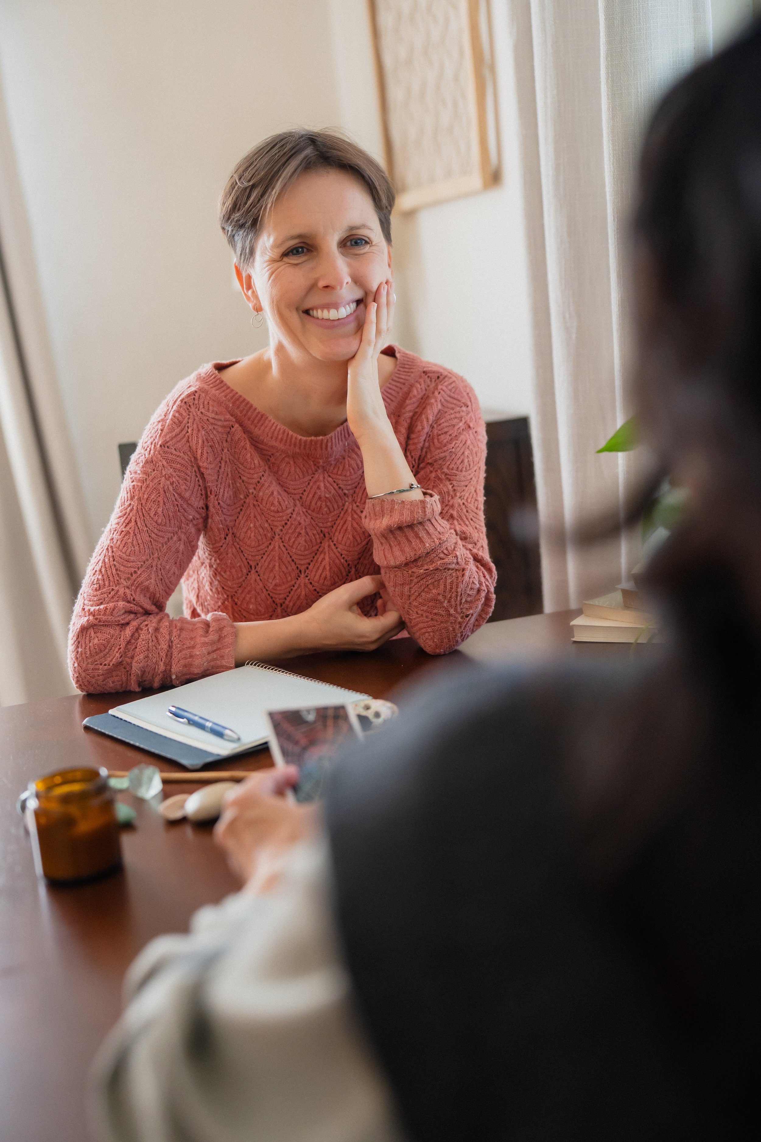 A woman with short brown hair and a pink sweater smiling while sitting at a table during a conversation.