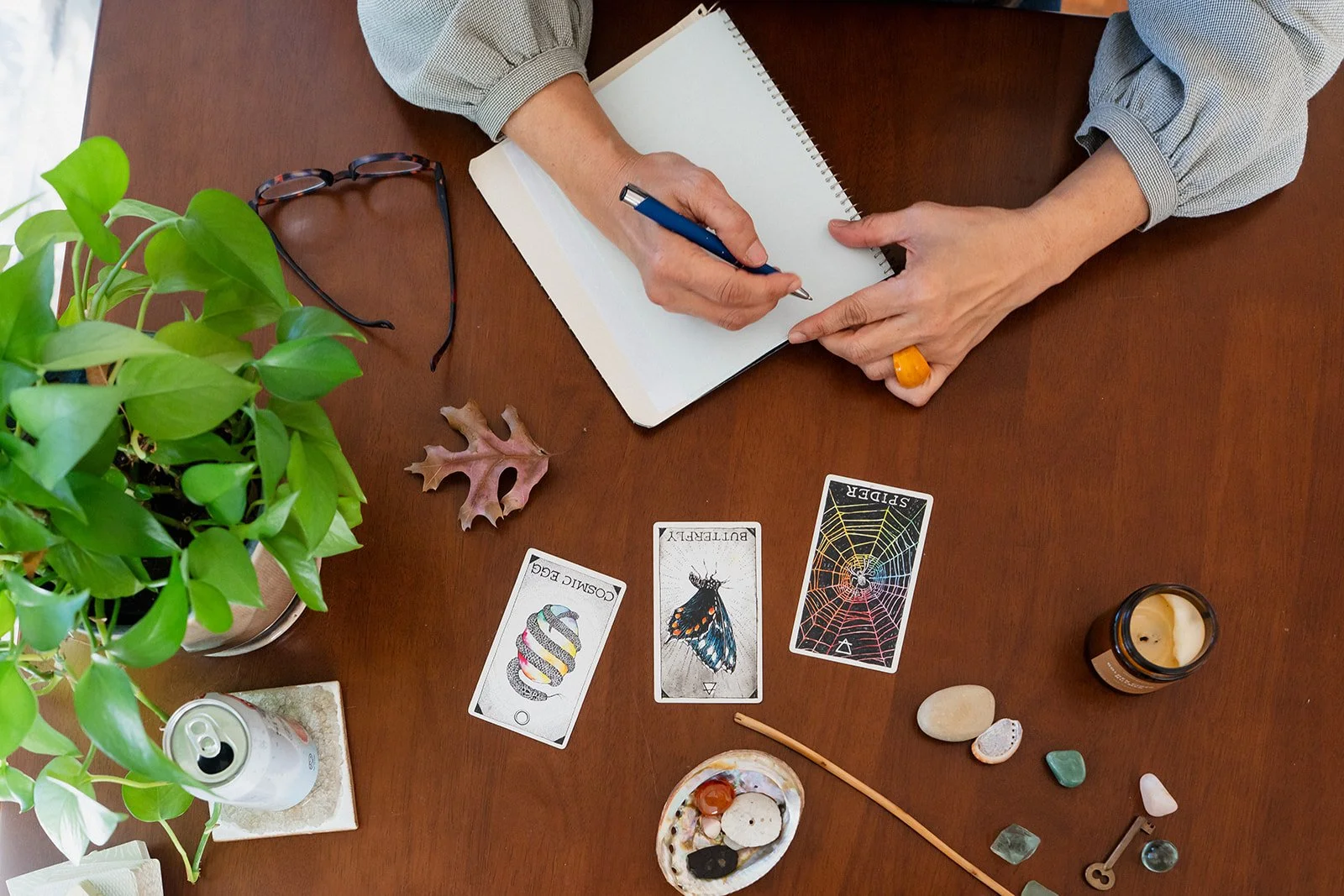 A person sitting at a wooden table with tarot and oracle cards, a notebook, glasses, a plant, rocks, a candle, and other small objects.