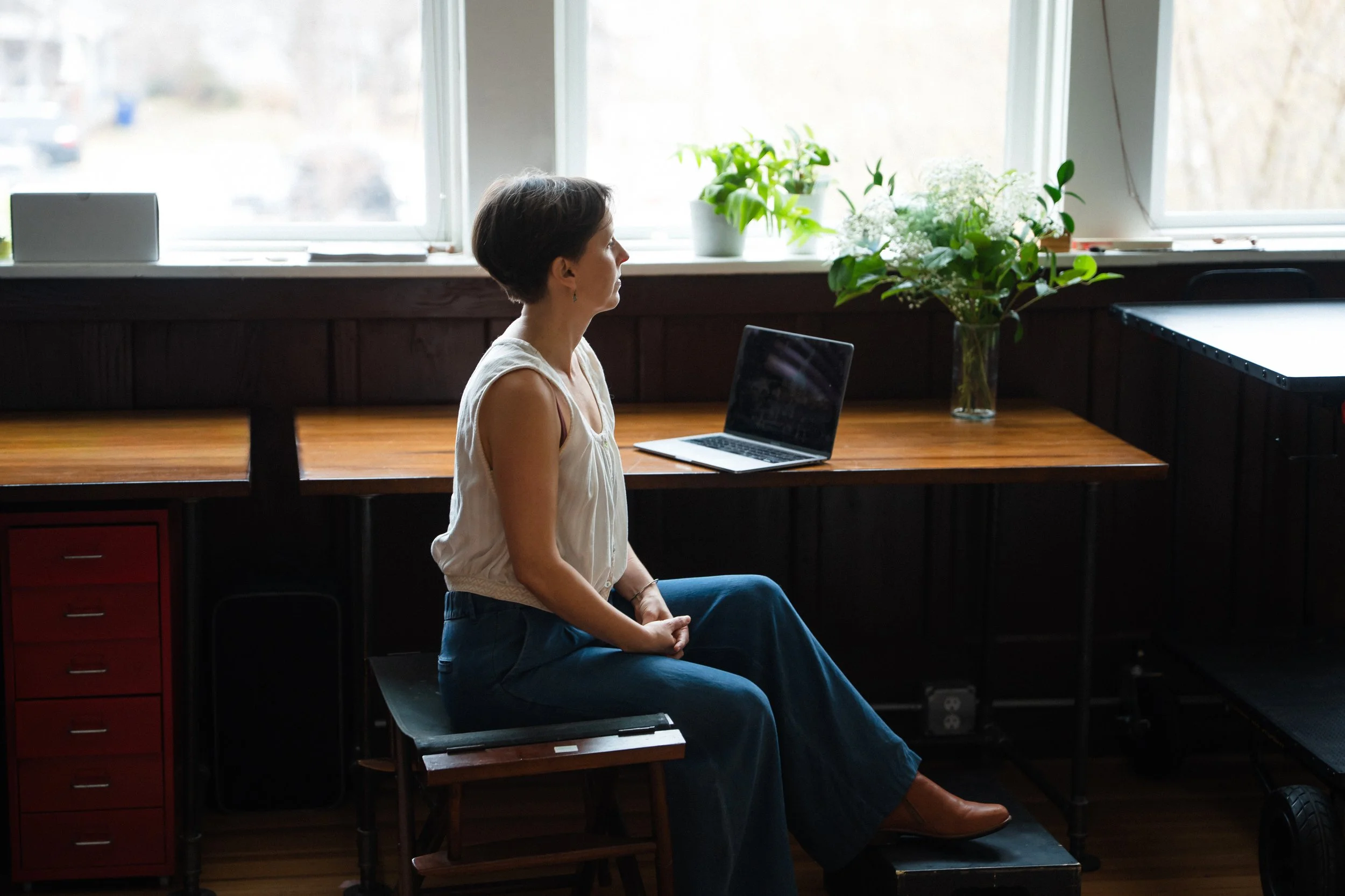 A woman sitting on a chair in front of a window with potted plants, a laptop, and a red cabinet in a cozy indoor space.