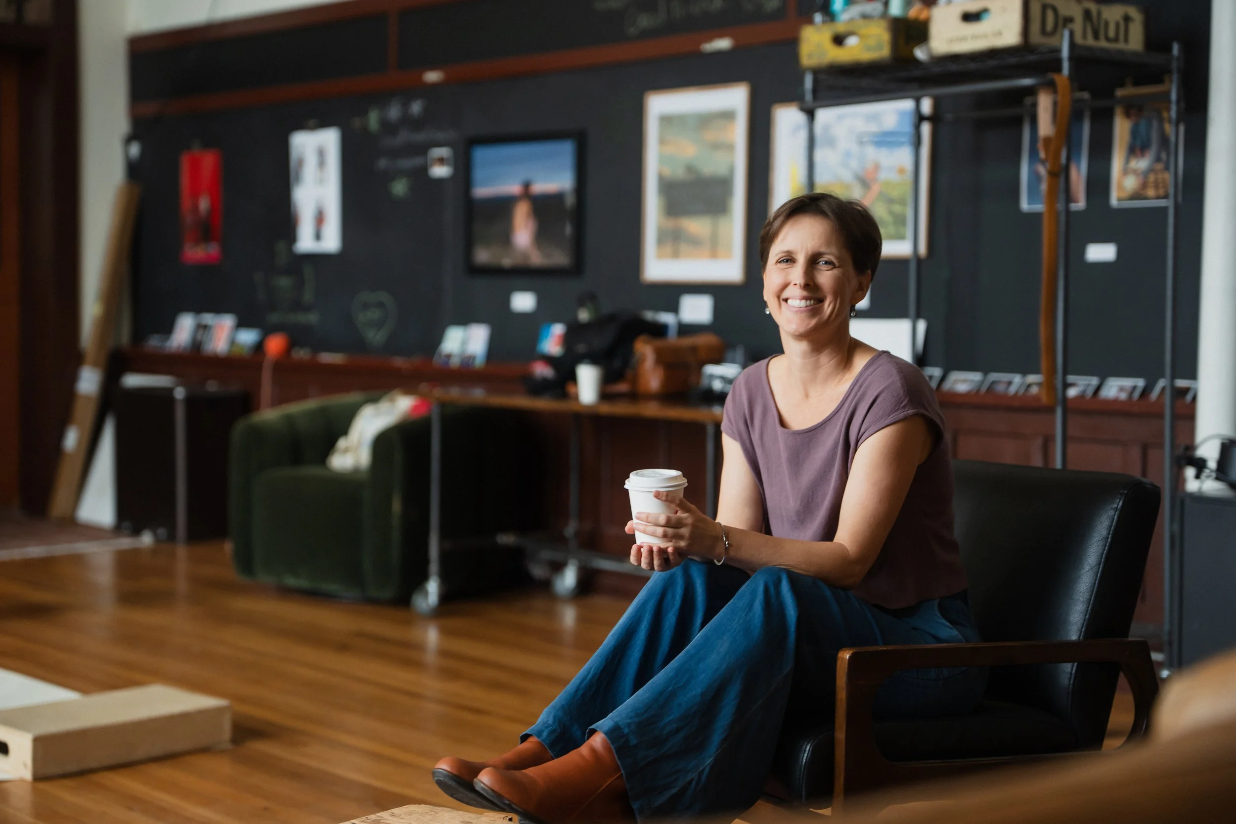 A woman smiling and sitting in a chair inside a room holding a cup, with artwork and photos on the black wall behind her.