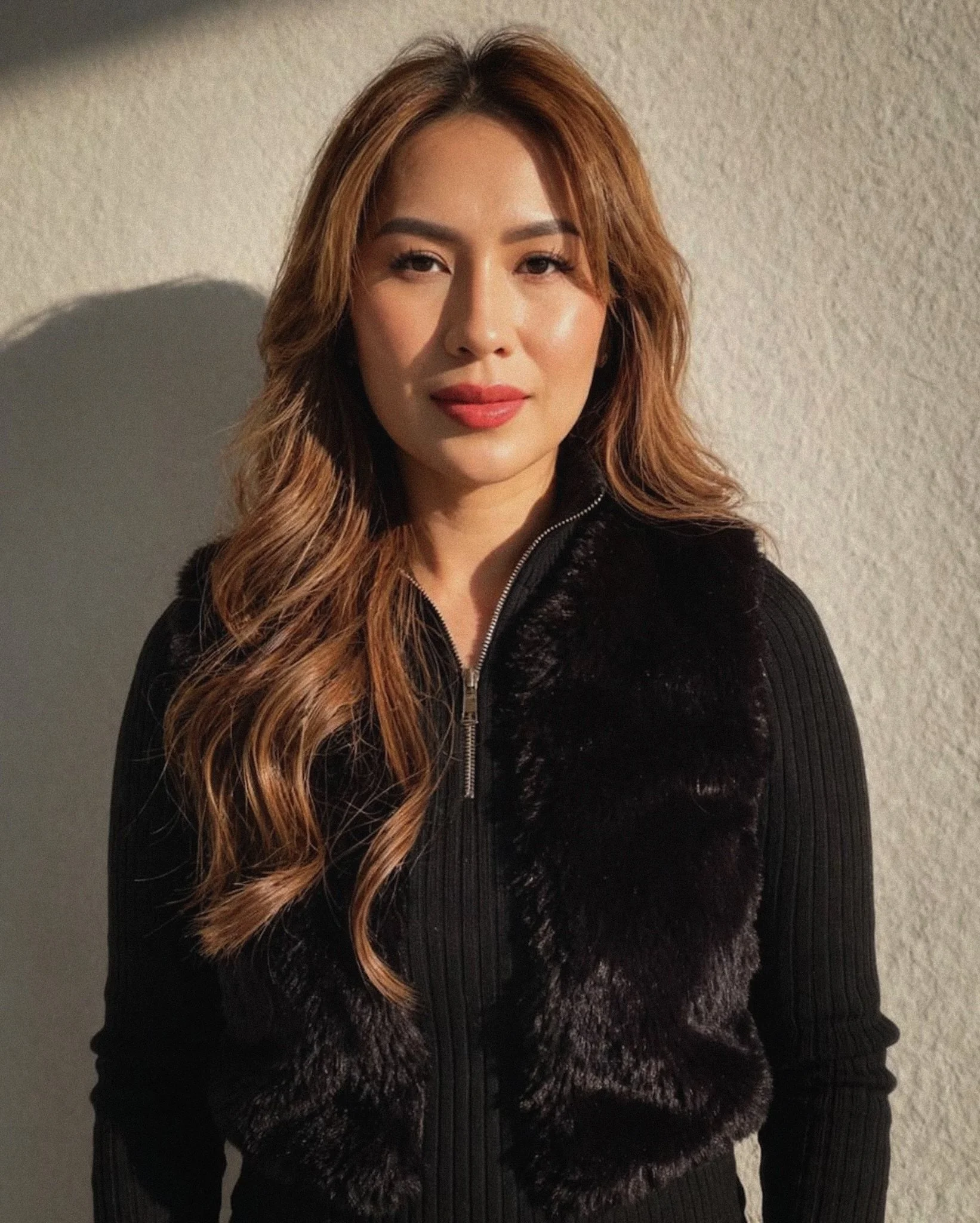 A woman with long wavy light brown hair, wearing makeup, a black zip-up sweater, and a black coat with faux fur trim, standing against a neutral-colored textured wall.