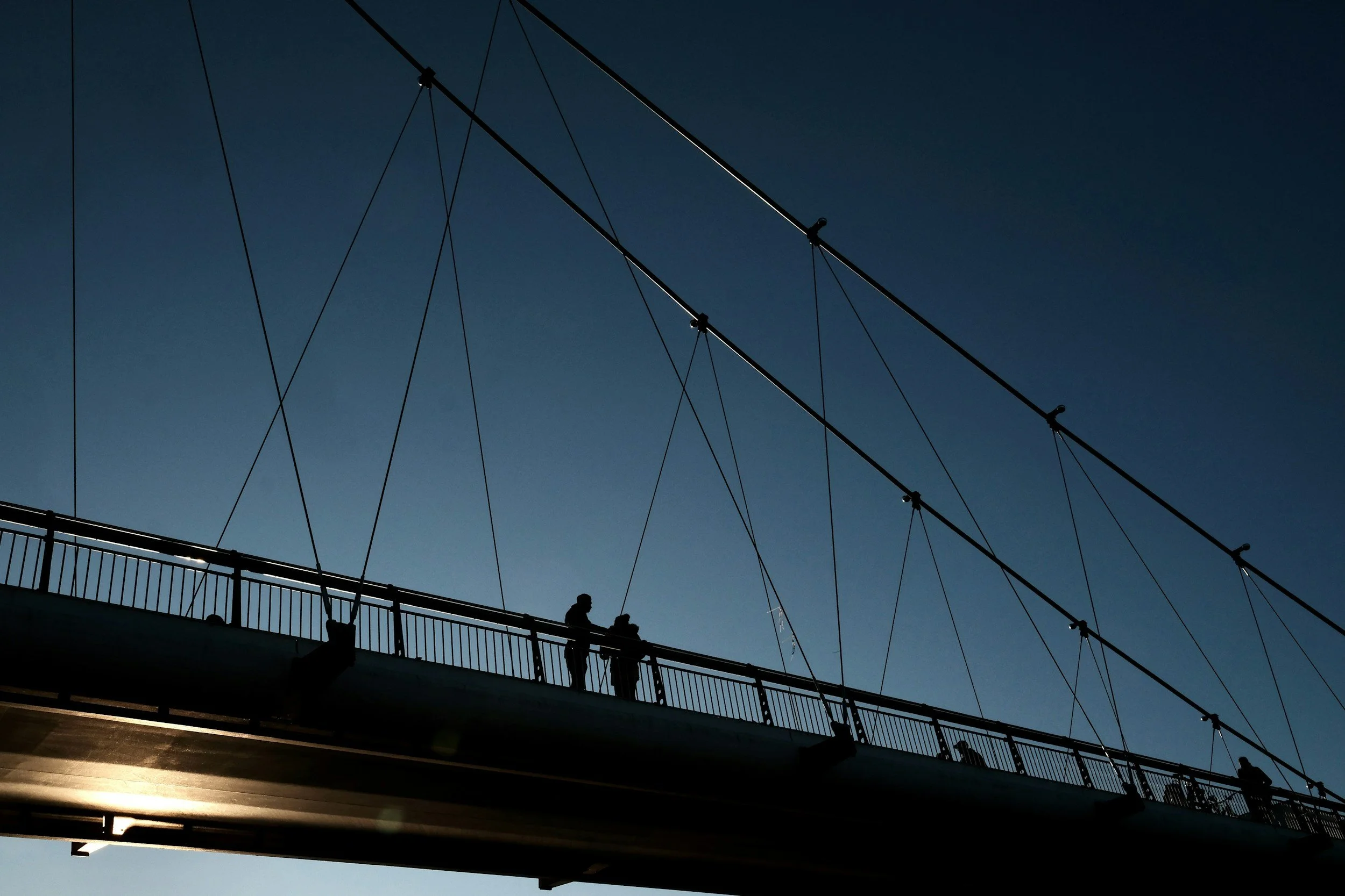 Silhouettes of people walking on a bridge with support cables against a clear blue sky.