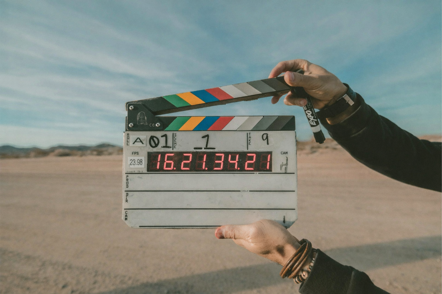 A person holding a film slate in a desert landscape under a blue sky with wispy clouds.