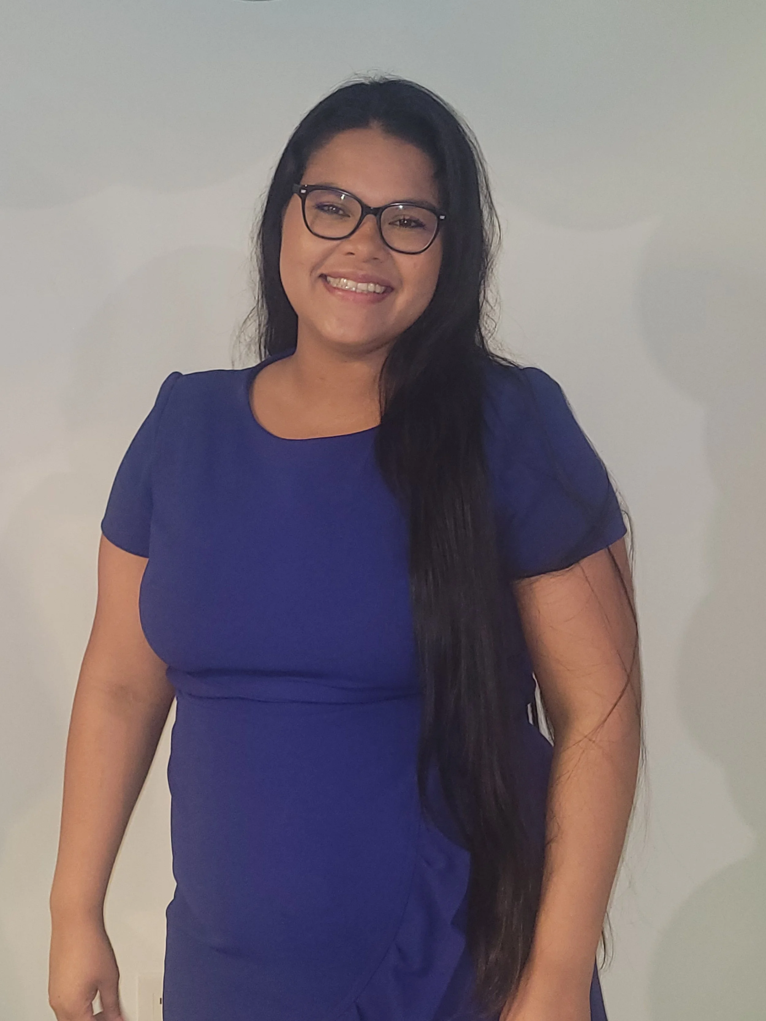A woman with long black hair, glasses, and a broad smile wearing a royal blue dress standing against a plain light-colored background.