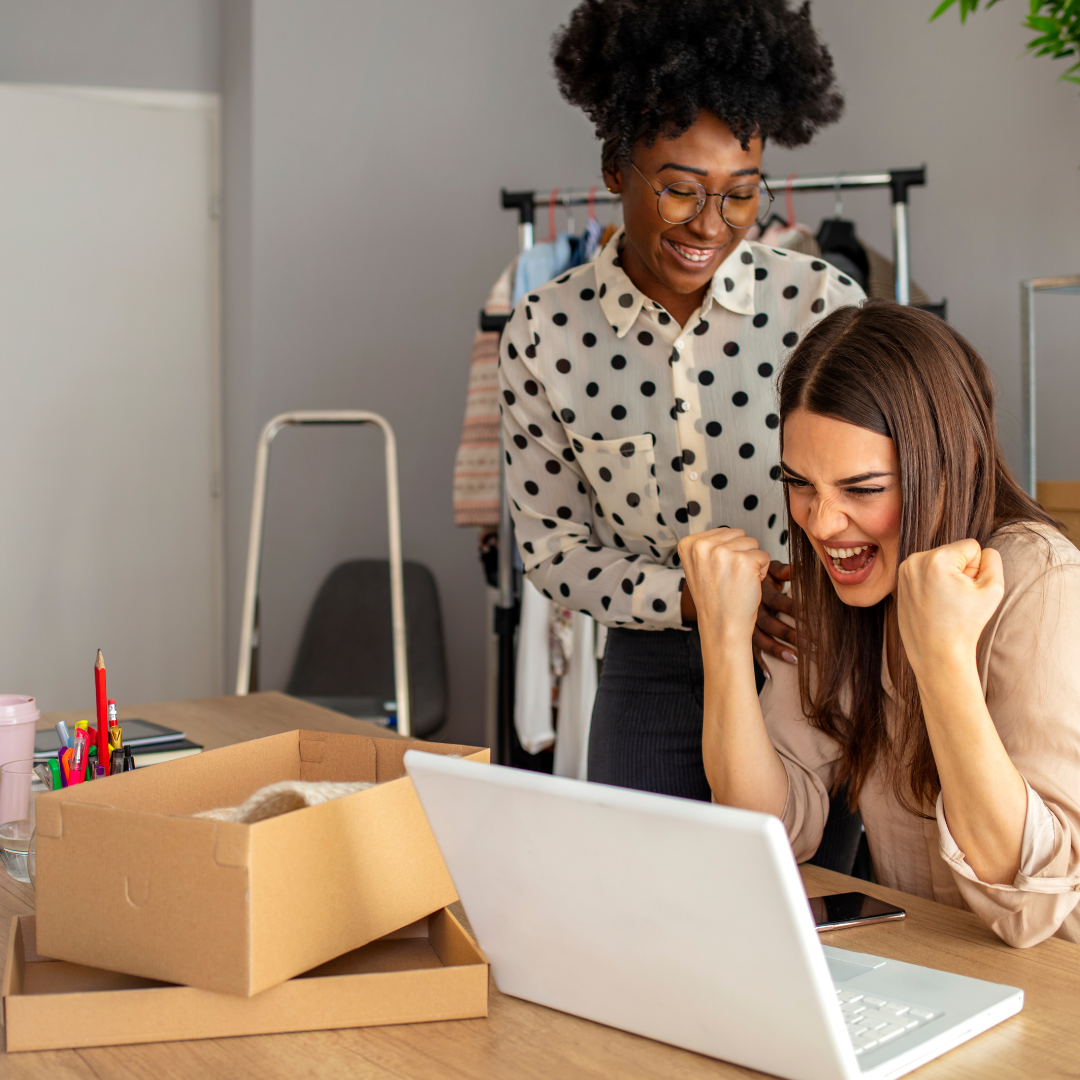 Two women celebrating success or achievement at a desk with a laptop, surrounded by office supplies and boxes, in a casual office setting.